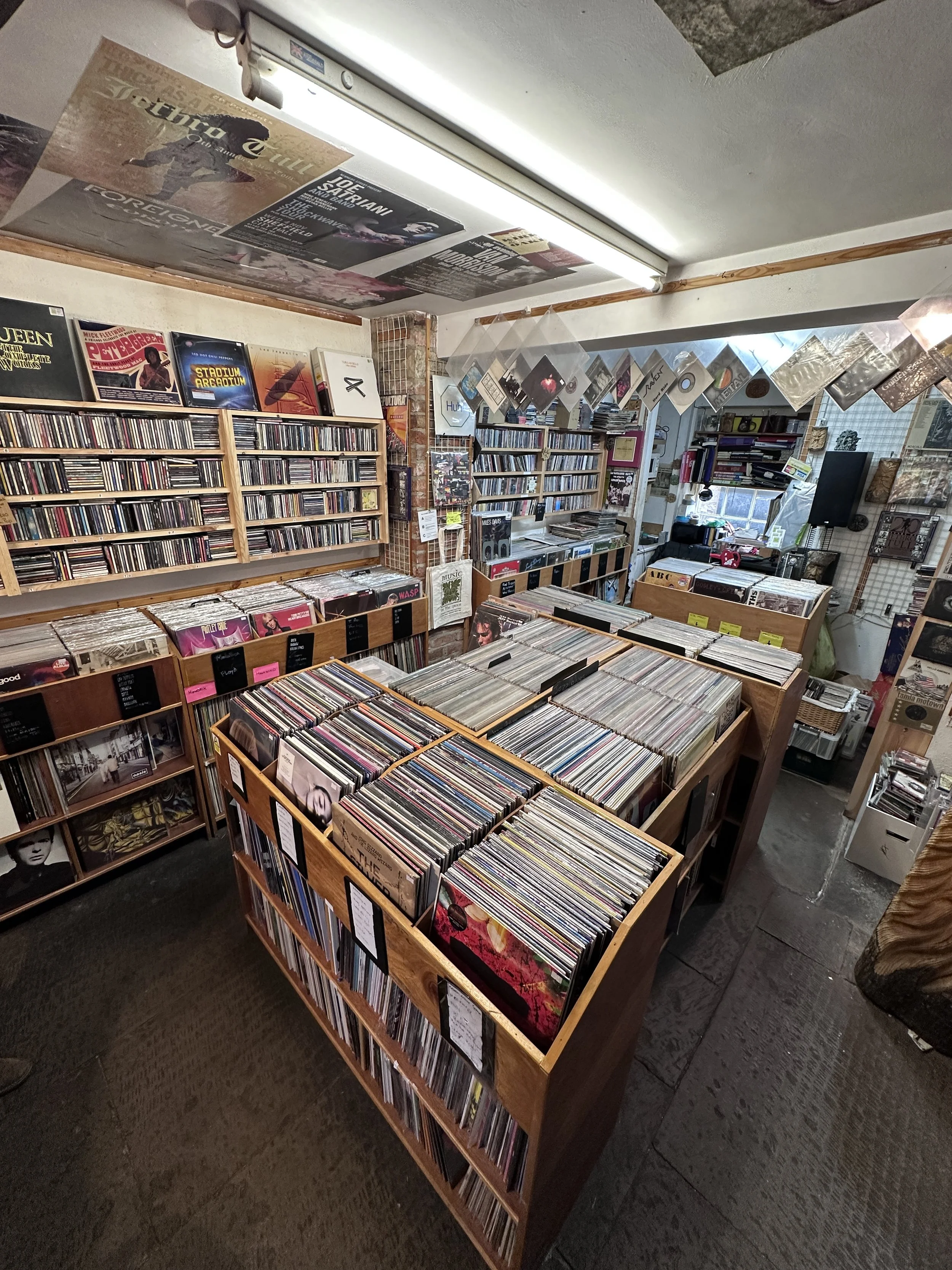 Interior of Music in the Green's Bakewell shop with wooden browser boxes filled with vinyl records. There are CDs on the racks and gig posters on the ceiling - the interior has a traditional record shop feel.