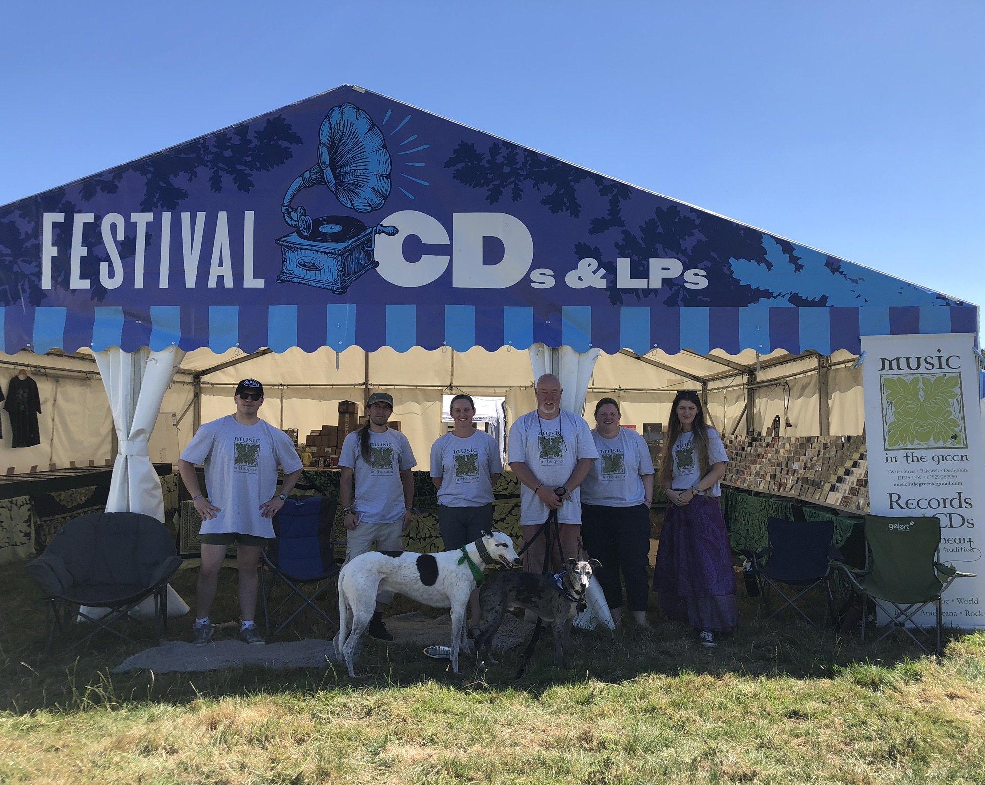 Music in the Green staff standing in front of their huge vinyl and CD marquee at Fairport Convention's Cropredy Festival. They are all wearing matching branded t-shirts with our familar green man logo and are accompanied by the owners two dogs. 