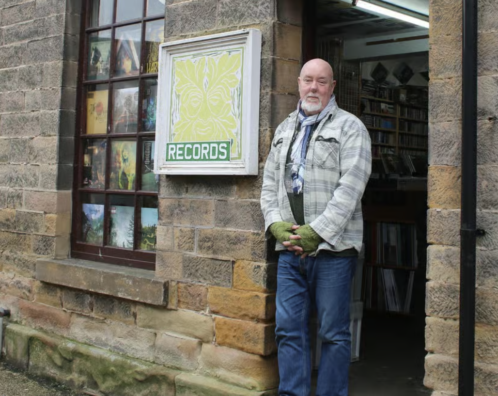 A man standing outside a record store with a sign that says 'RECORDS'. He is wearing a plaid jacket, jeans, fingerless gloves, and a scarf, and is standing beside a large window on a brick building.