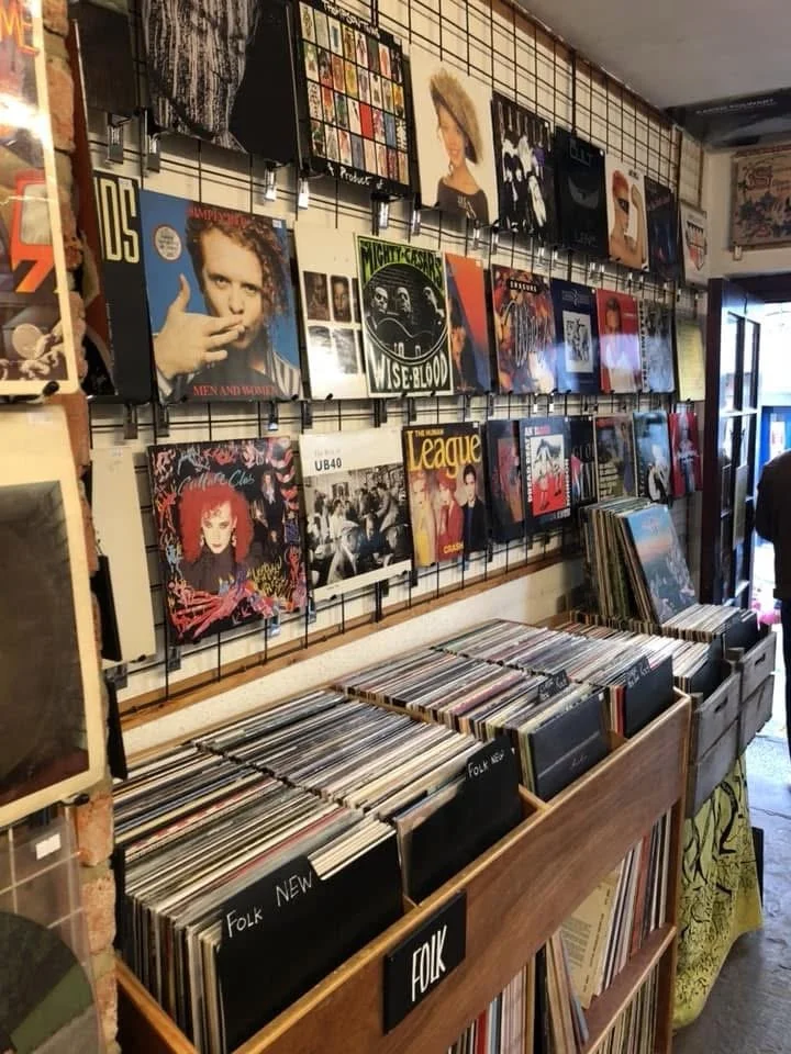 Red-haired woman with makeup on music album cover, among other vinyl records and album covers on display at a record shop.