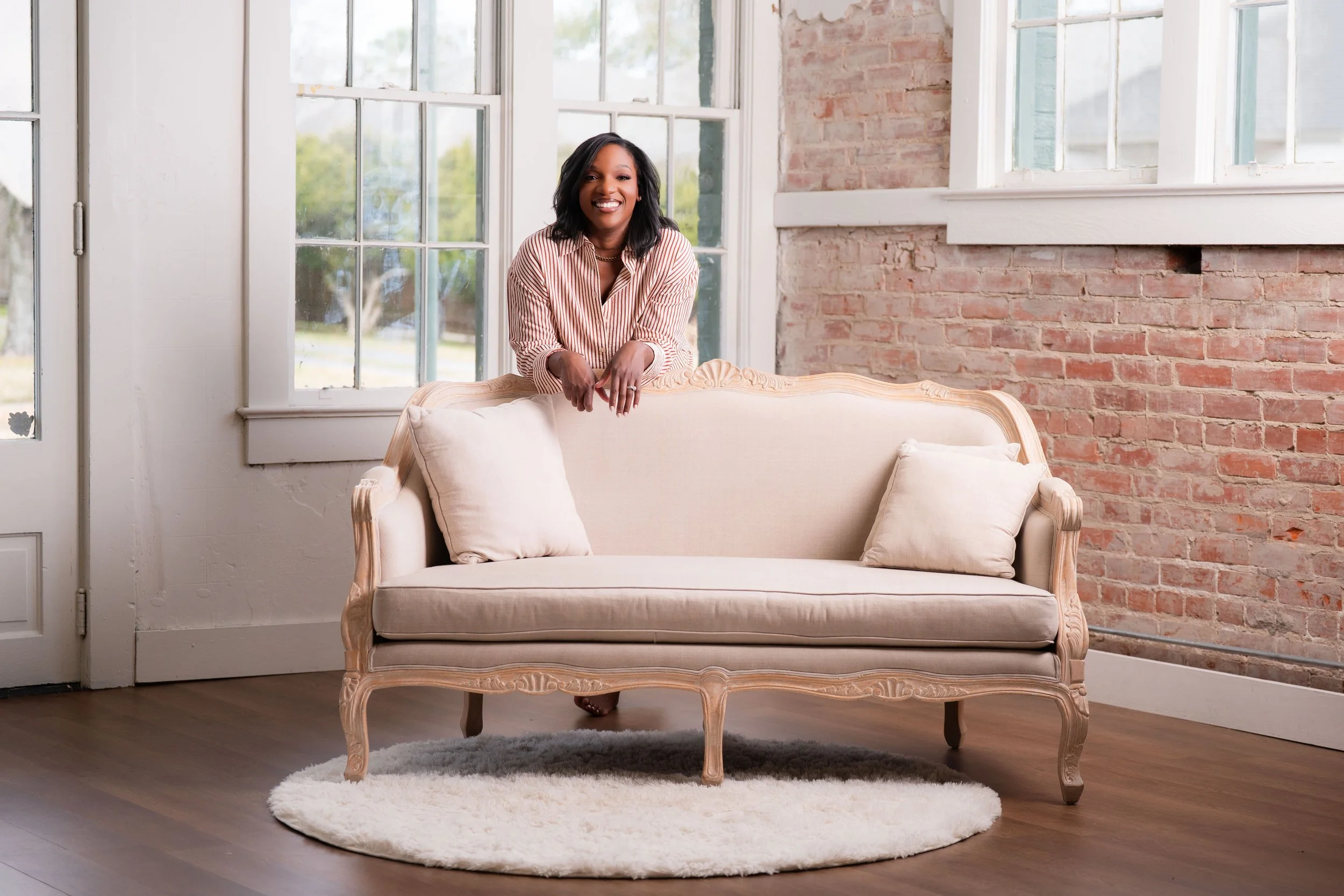 A woman with dark hair and a striped shirt smiling while leaning on a vintage cream-colored sofa inside a room with brick and white walls and large windows.