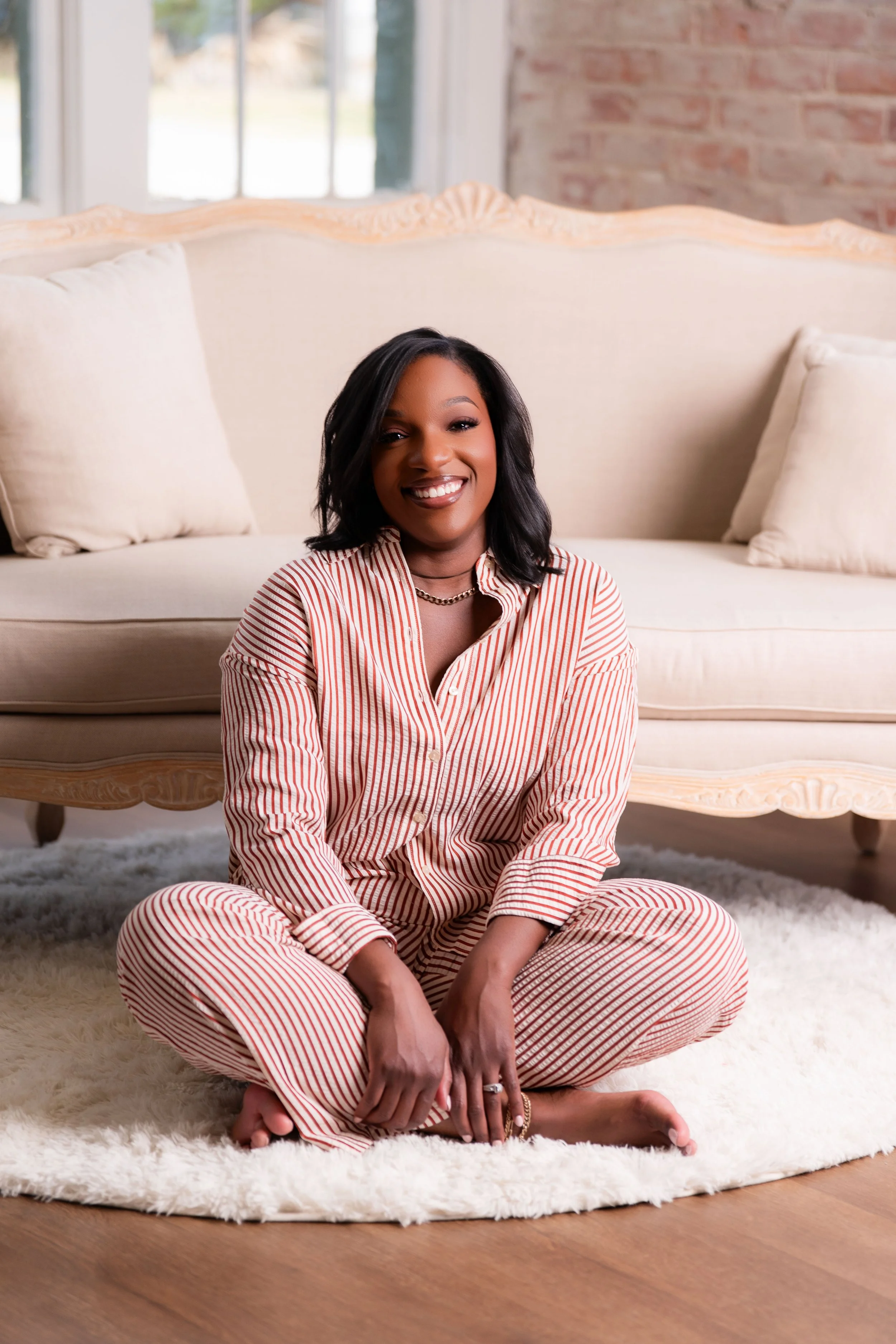 A woman with dark hair and medium skin tone sitting on a fluffy white rug in front of a beige sofa with matching pillows, wearing red and white striped pajamas, smiling at the camera, in a well-lit room with large windows and a brick wall.