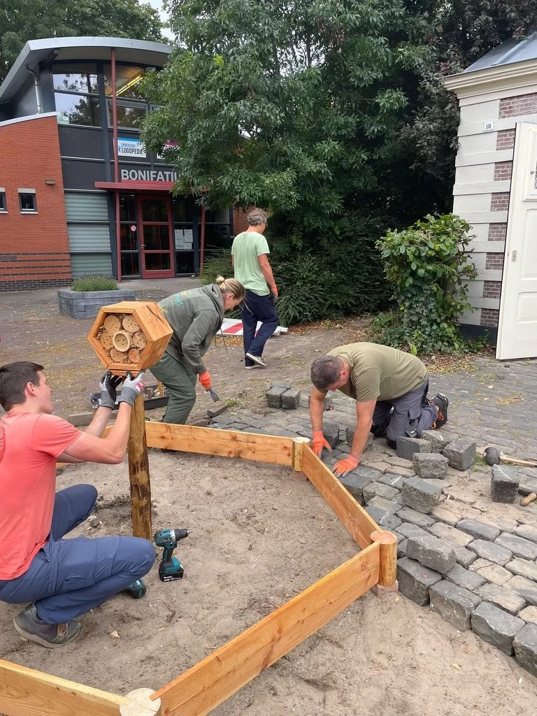 Mensen leggen bestratingsstenen rondom een Beewell-tuin, terwijl één persoon een bijenhotel vasthoudt.