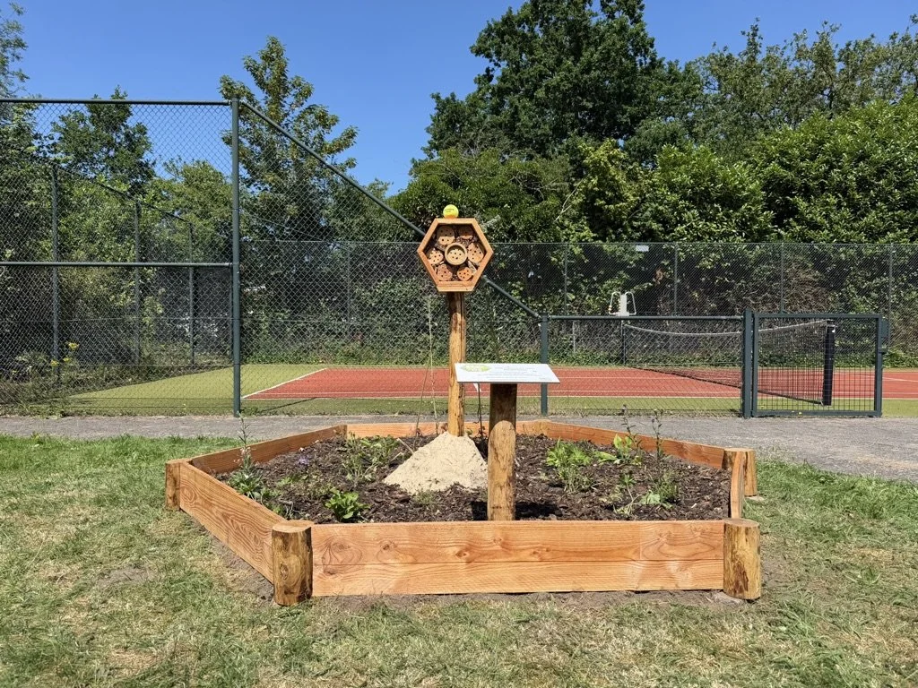 Een Beewell-tuin met bijenhotel op een veld met tennisbanen op de achtergrond.