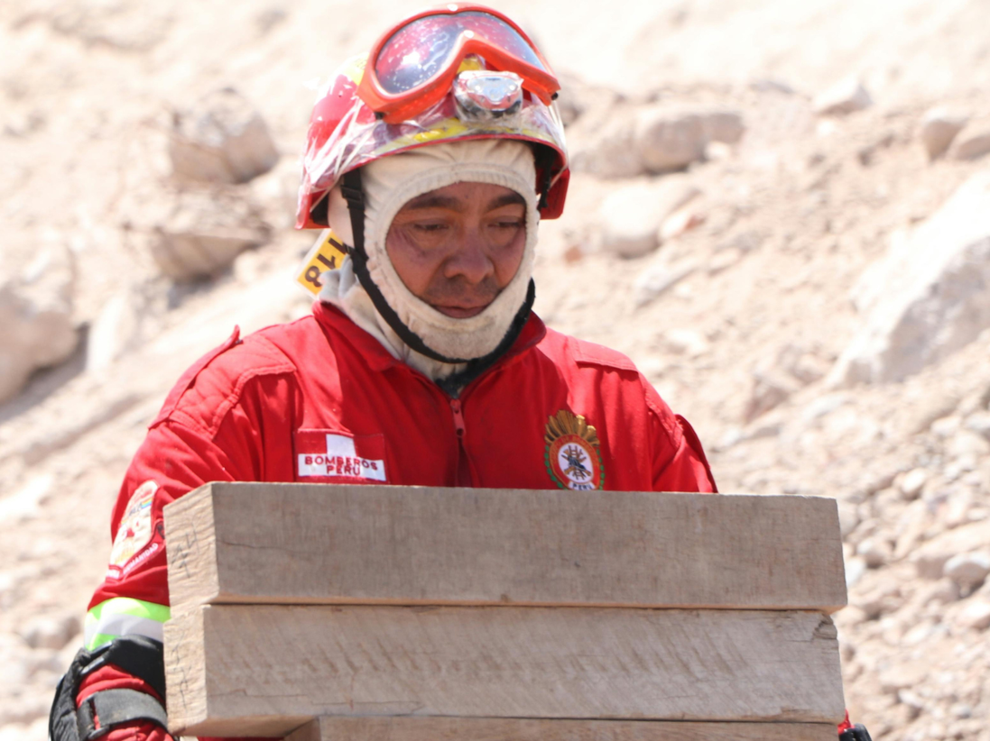 An aid worker wearing red boiler suit helmet and goggles, moves a stack of timber.
