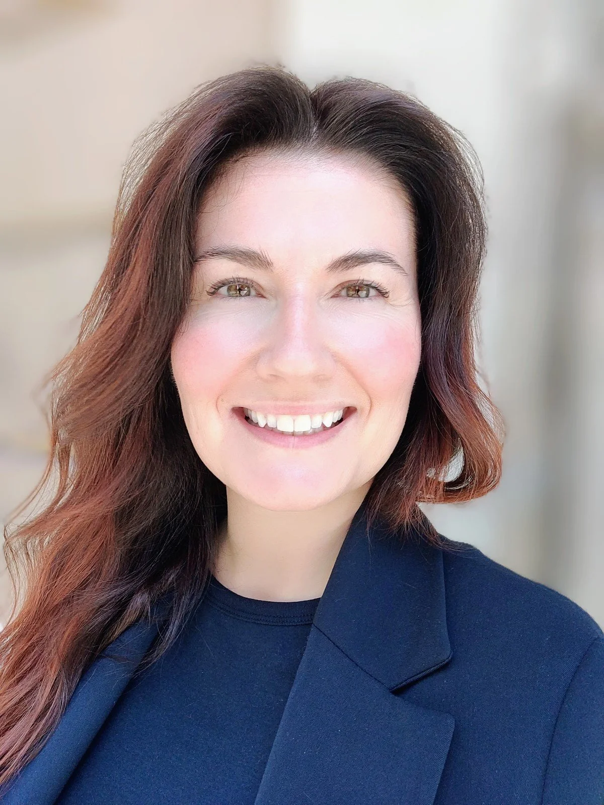 A smiling woman with shoulder-length wavy brown hair wearing a navy blazer and a black shirt, standing outdoors with a blurred neutral background.