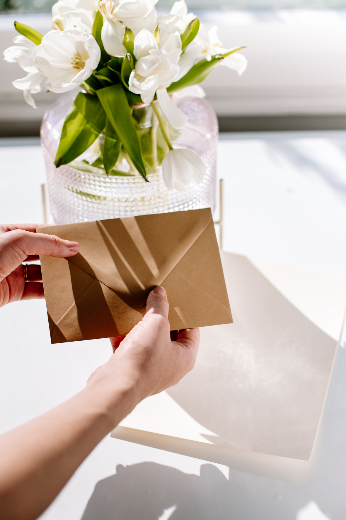 Person opening a brown envelope in front of a glass vase with white tulips on a white surface, near a window with sunlight casting shadows.