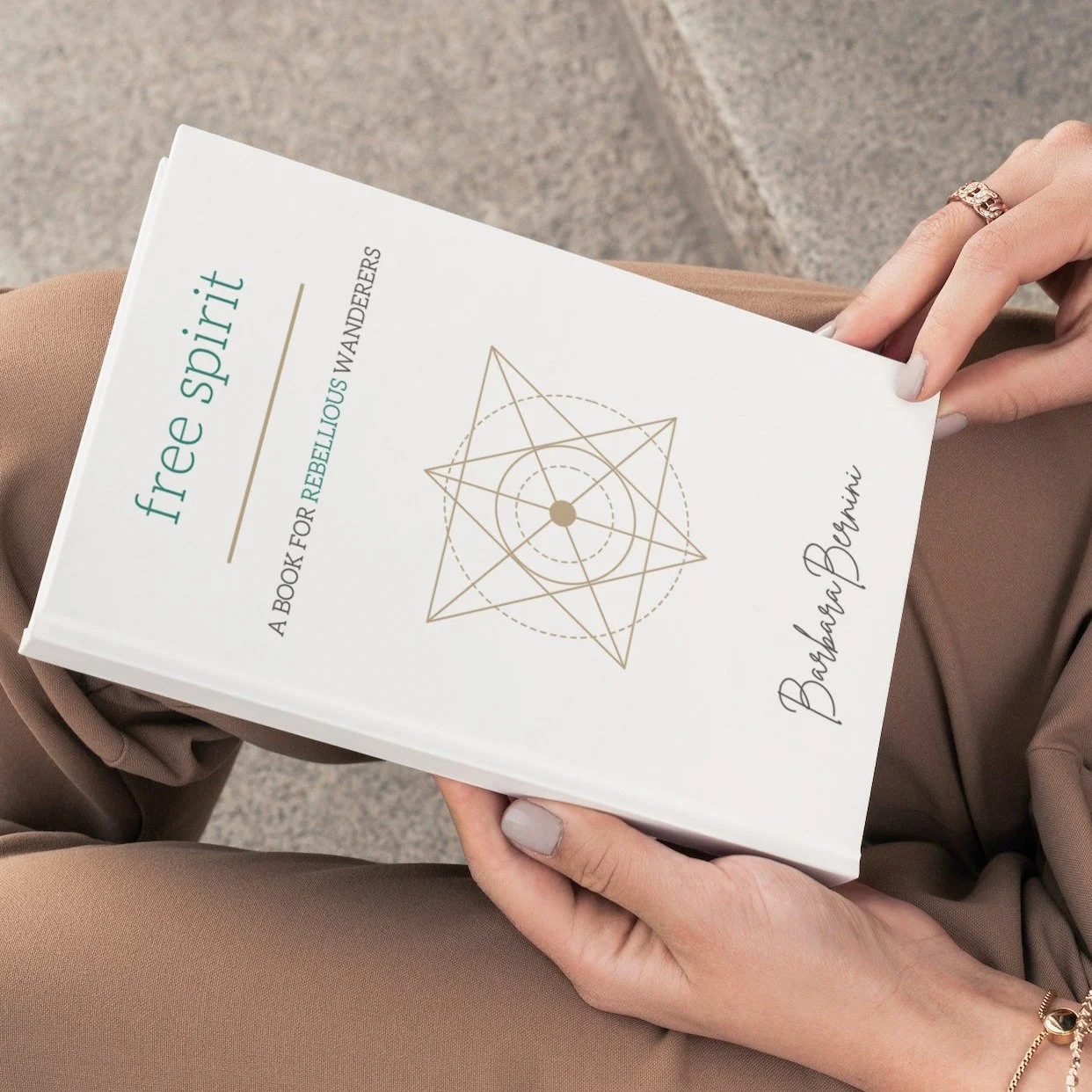 Close-up of a person holding a book titled "free spirit: A Book for Rebellious Wanderers" by Barbara Bonini, sitting outdoors on numbered stone steps.