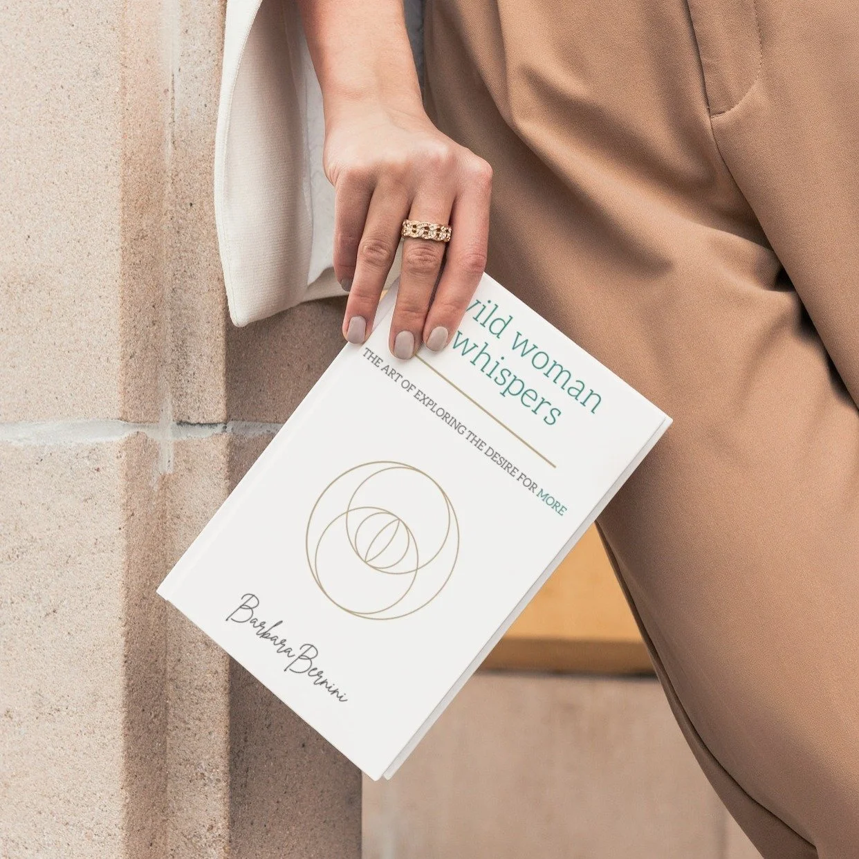 A woman holding a book titled "Wild Woman Whispers: The Art of Exploring the Desire for More" with her left hand, wearing a gold ring on her ring finger, and dressed in beige pants and a white blazer, standing against a brick wall.