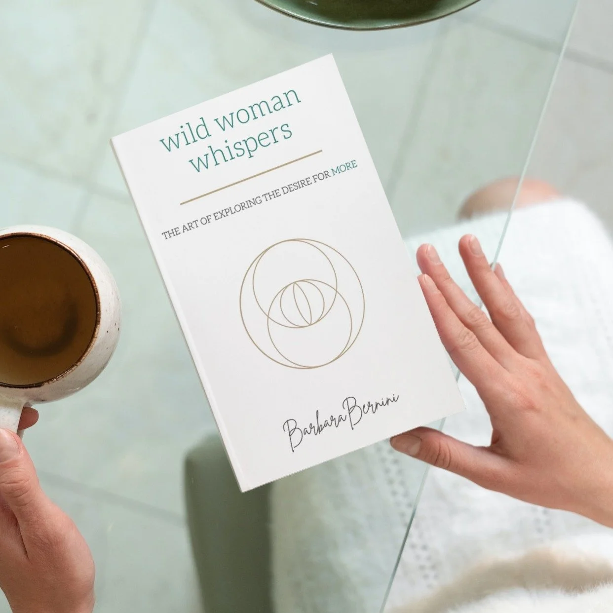 A woman holding a coffee mug in one hand and a book titled 'Wild Woman Whispers' by Barbara Bernini in the other, standing near a glass table.