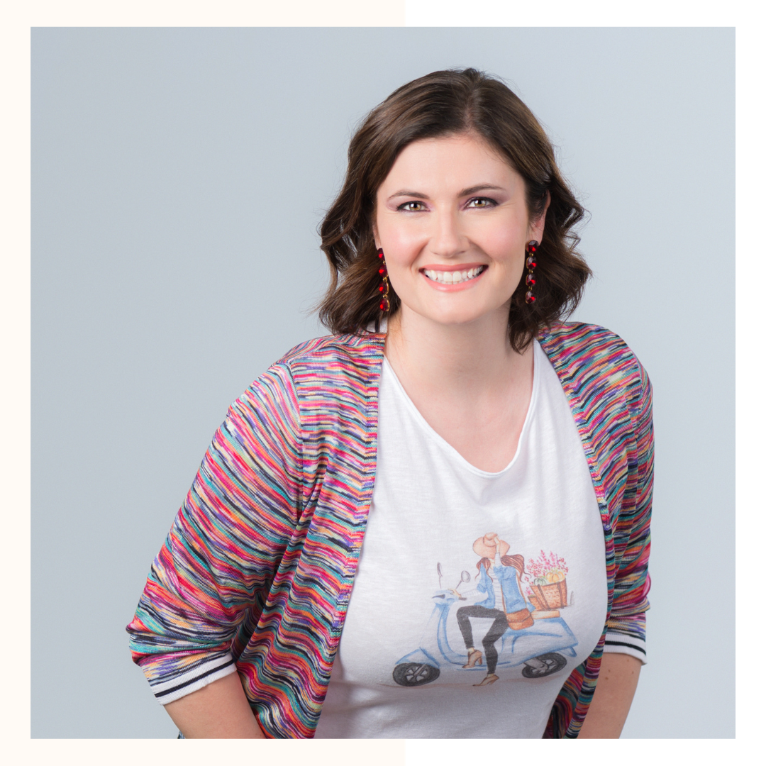 A woman with shoulder-length brown hair smiling, wearing red earrings, a white t-shirt with a graphic of a woman on a scooter, and a colorful striped cardigan against a plain light gray background.