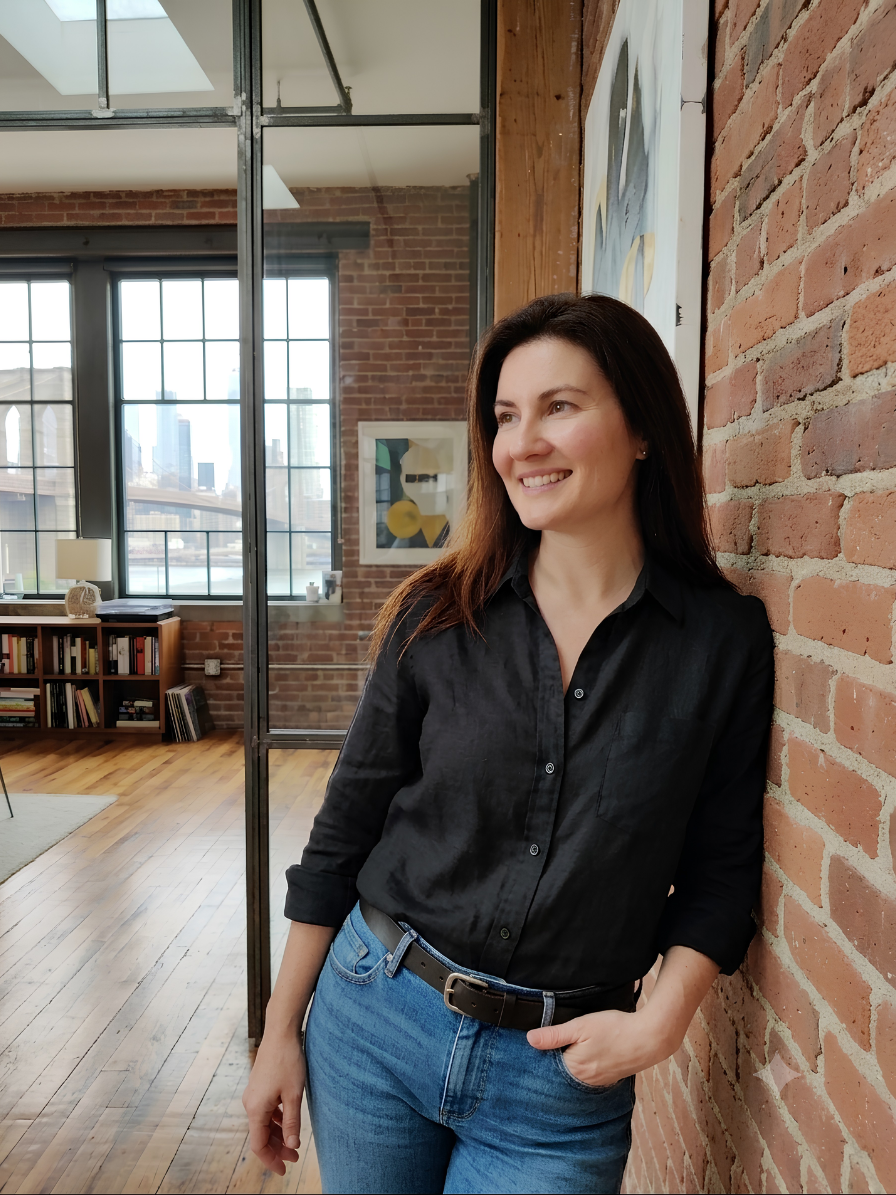 A woman with long brown hair smiling, leaning against a brick wall in a modern loft-style apartment with large windows, wooden floors, and an art piece on the wall.