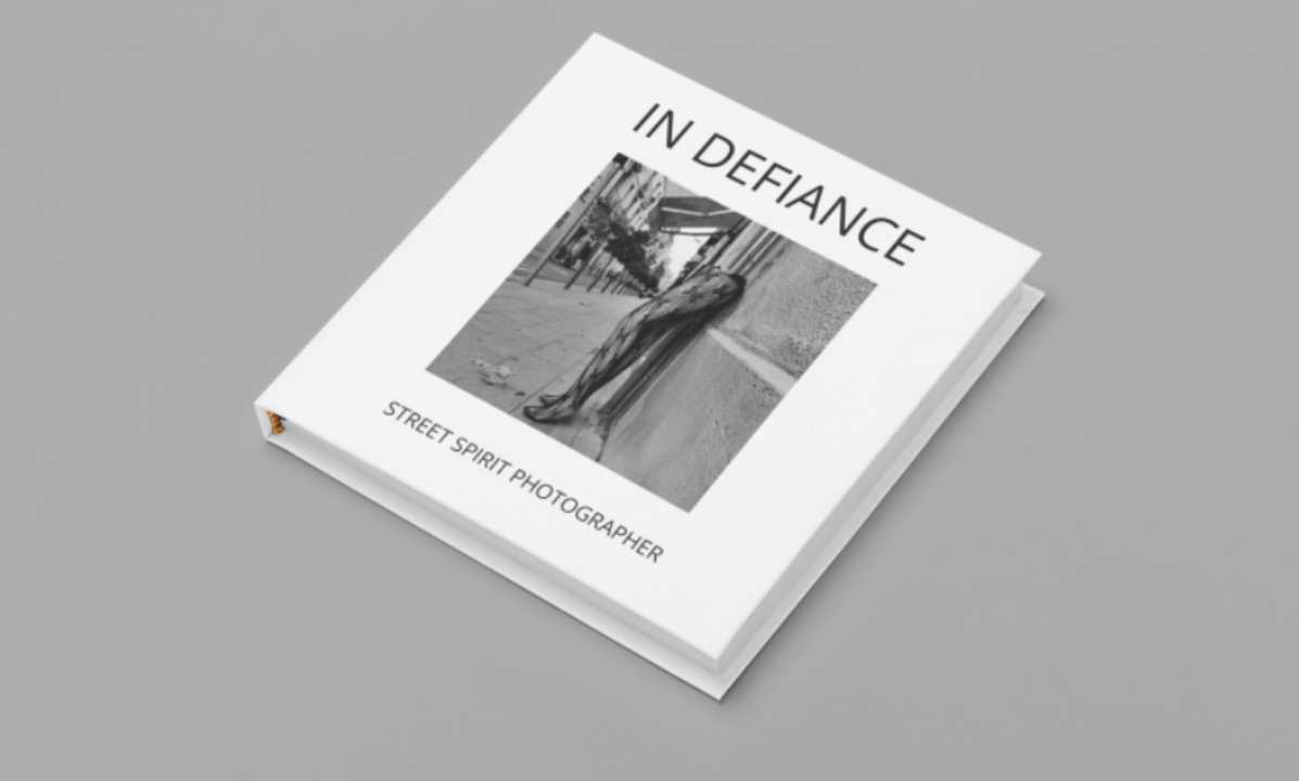 A book titled "In Defiance" with a black and white cover photo of a street scene featuring a person walking on the sidewalk, and the subtitle "Street Spirit Photographer".