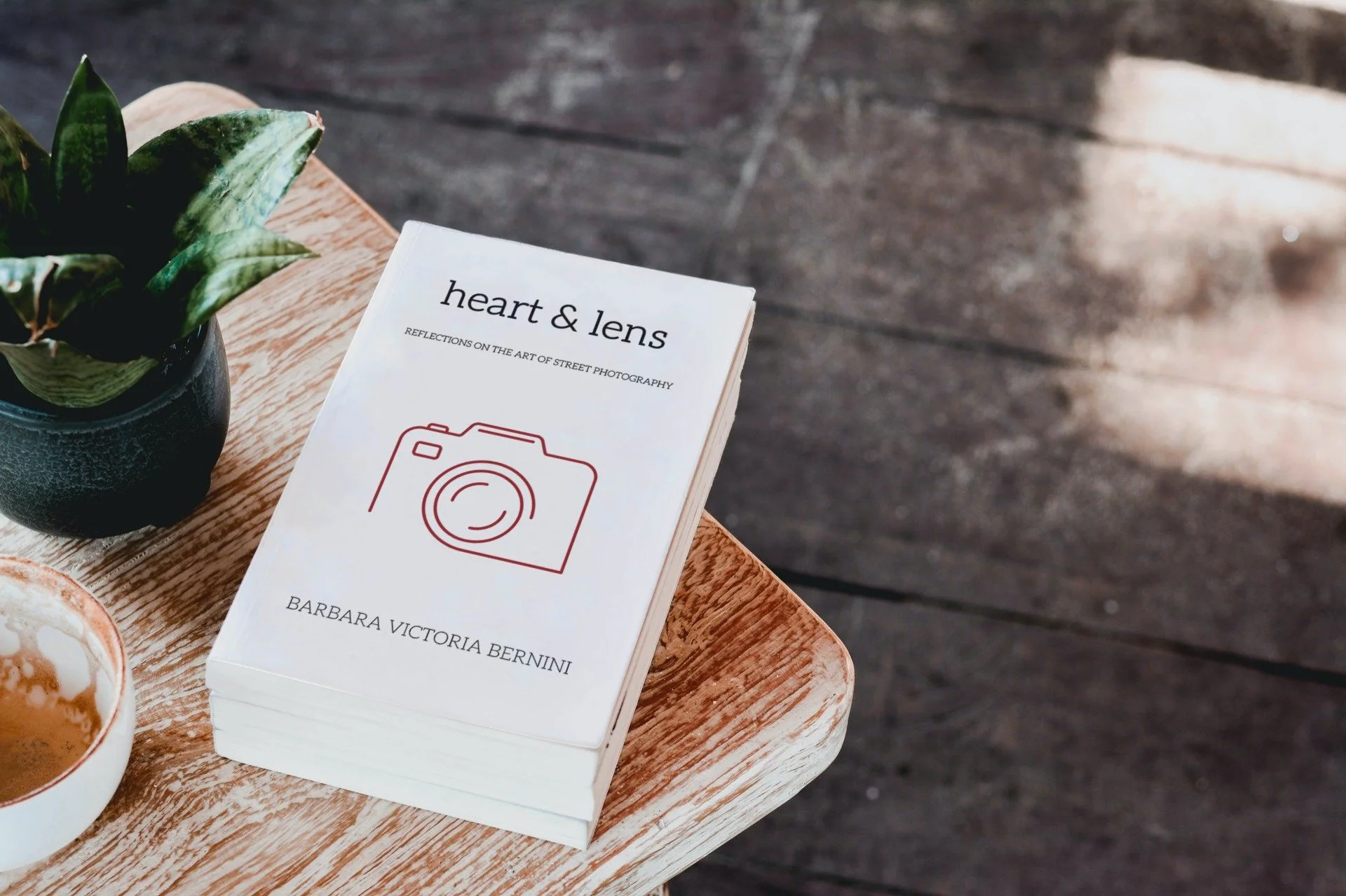 A book titled 'heart & lens' with a subtitle 'Reflections on the Art of Street Photography' rests on a wooden surface next to a potted plant and two cups of coffee on a wooden table.
