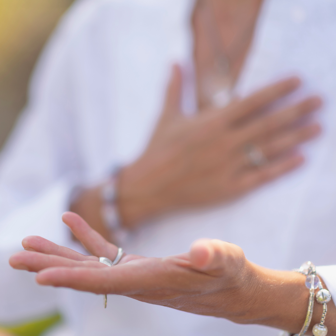 A woman with multiple rings and bracelets, wearing a white top, with her hand on her chest and another hand extended outward.