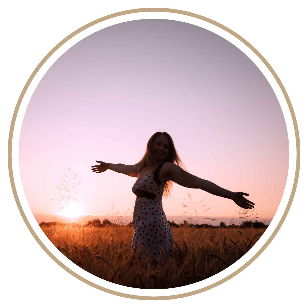 A woman standing in a field during sunset with outstretched arms, smiling, wearing a polka dot dress.