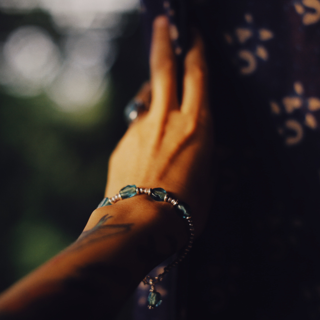 Close-up of a hand with a bracelet, resting on a dark surface with a pattern, with a blurred background.