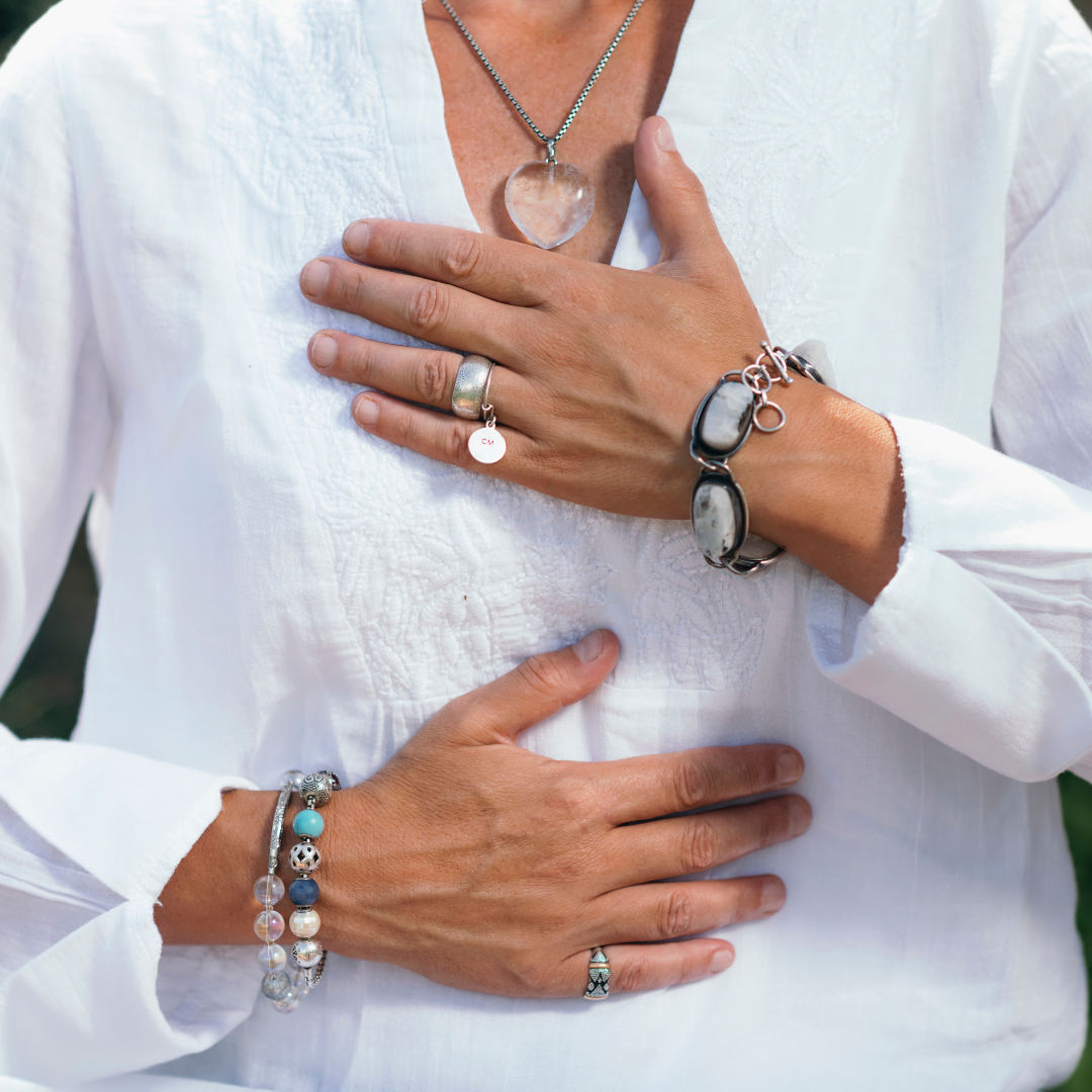 Two hands with jewelry placed on the chest and stomach of a woman wearing a white blouse, displaying necklaces, rings, bracelets, and a ring.