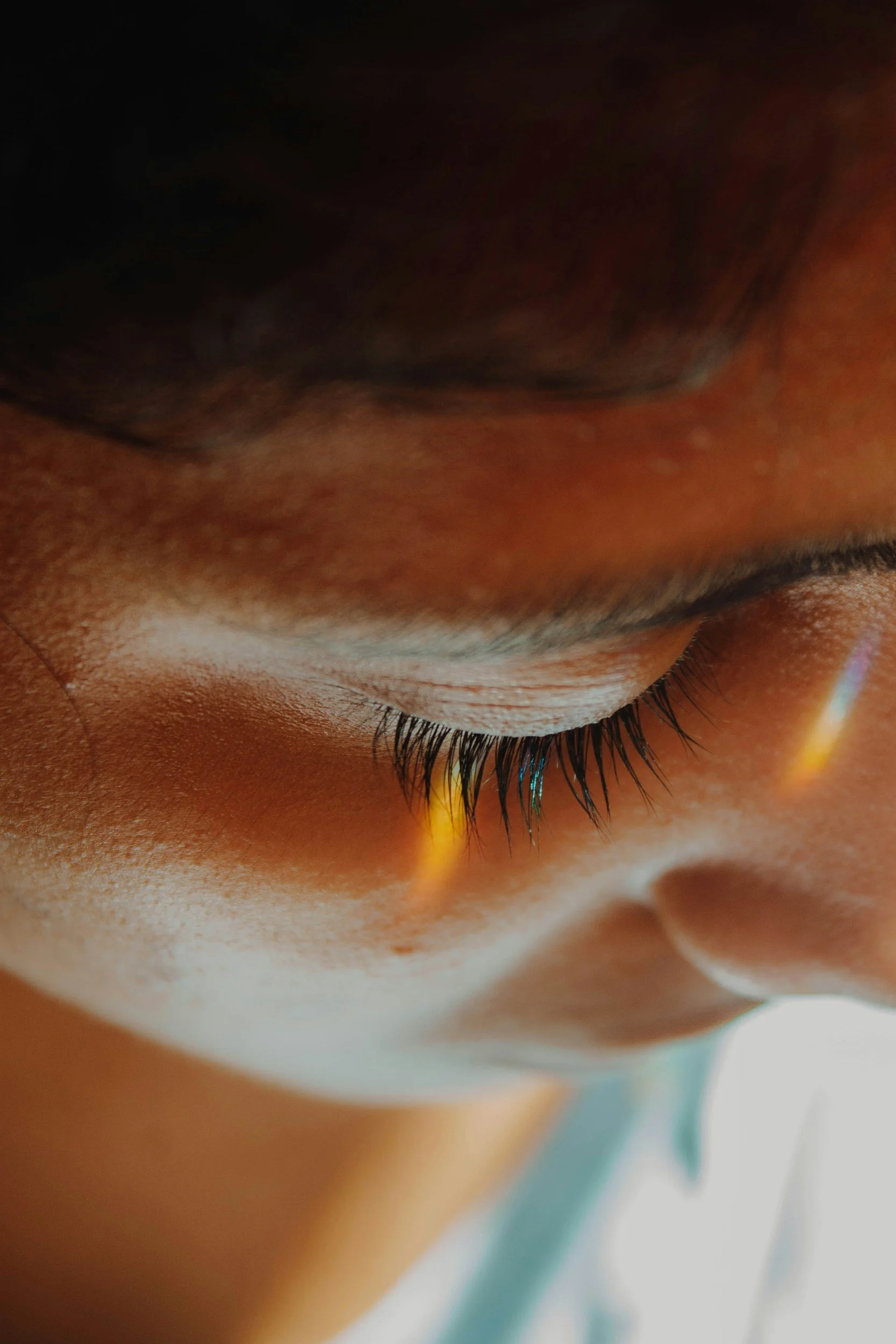 Close-up of a person's closed eye showing eyelashes, eyelid, and part of the face with warm lighting and a faint rainbow reflection.