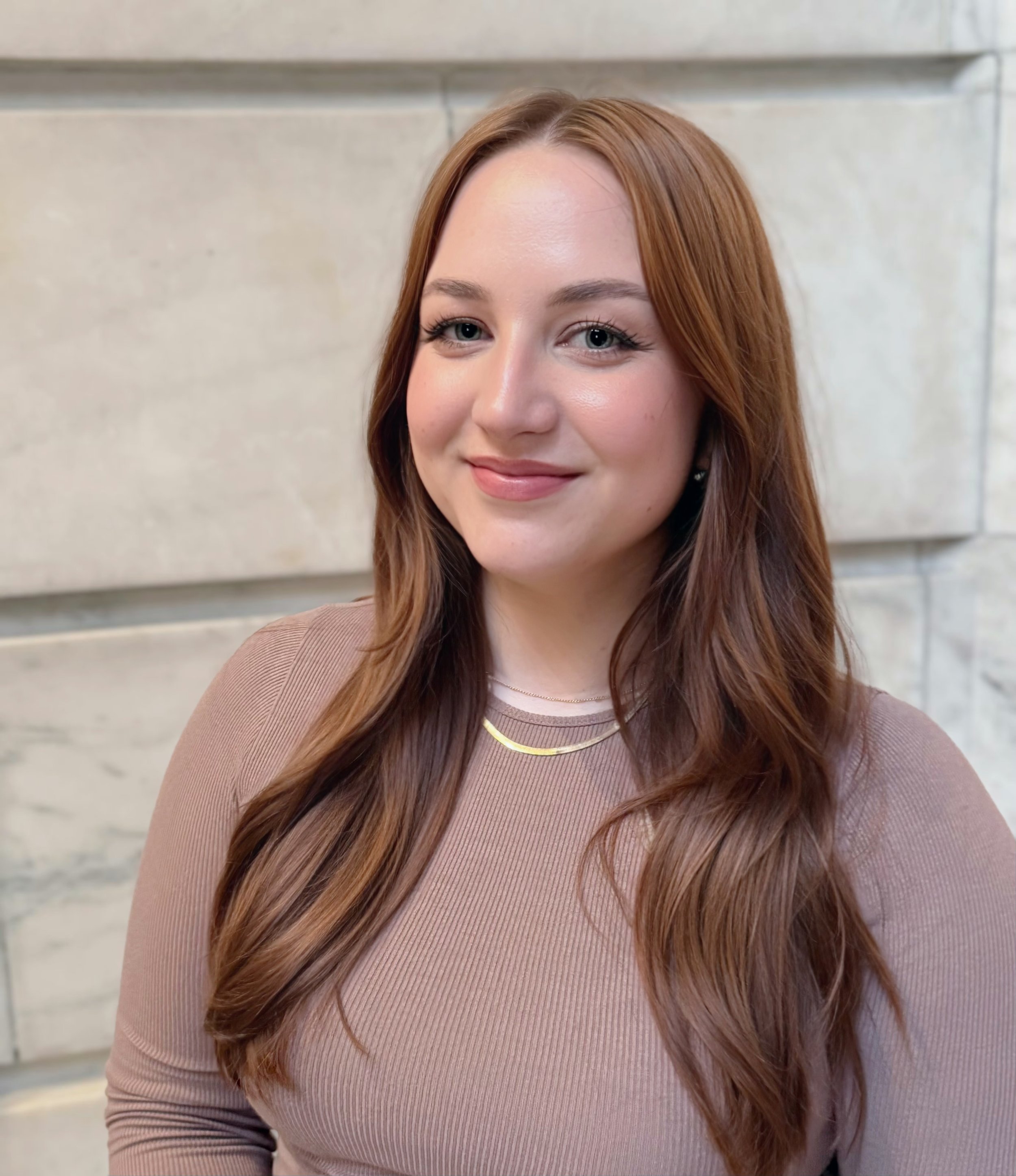 A young woman with long auburn hair and blue eyes smiling outdoors against a stone wall.