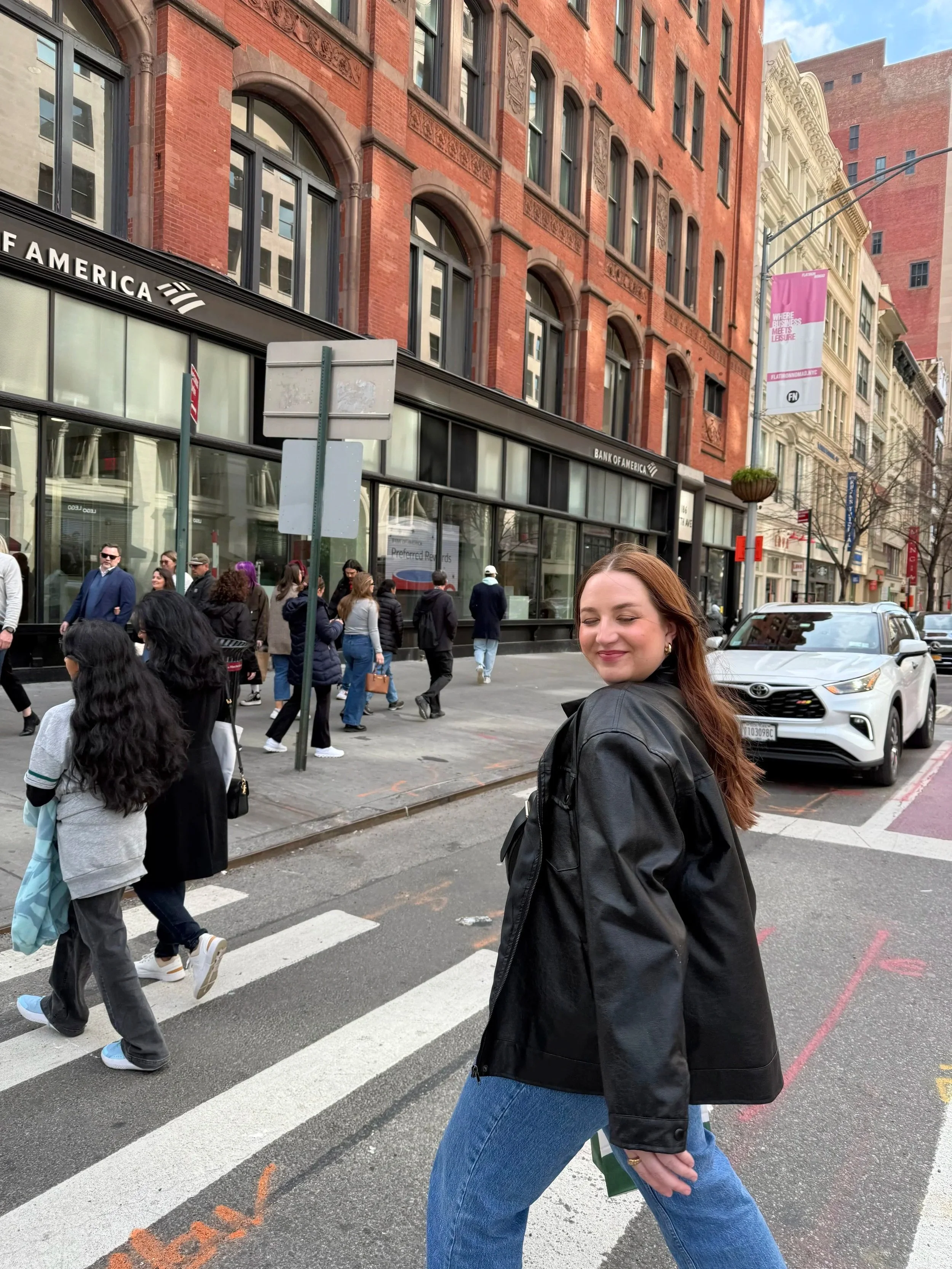 A woman with red hair wearing a black leather jacket and blue jeans crossing the street in New York City at a crosswalk in an urban area with brick buildings and a Bank of America branch in the background. Other pedestrians walk along the sidewalk.