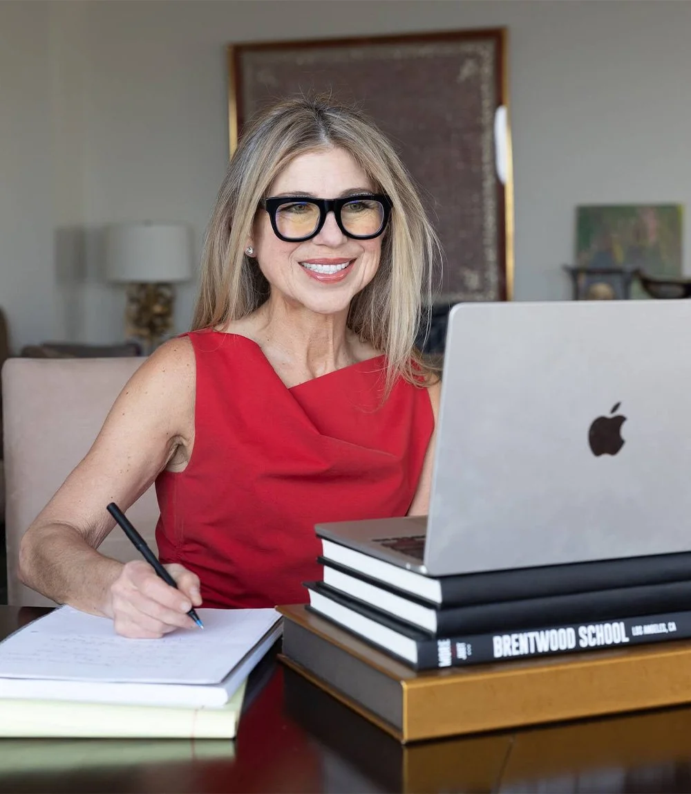 Professional woman in red dress and black glasses smiling at desk with laptop and notebook in modern home office