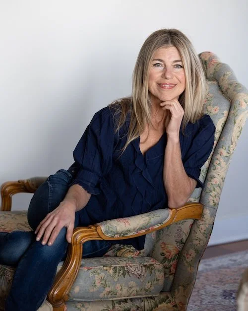 Smiling middle-aged woman with long blonde hair sits relaxed in a floral armchair, wearing a dark blue blouse and jeans, resting her chin on one hand and looking at the camera against a plain light-colored wall.