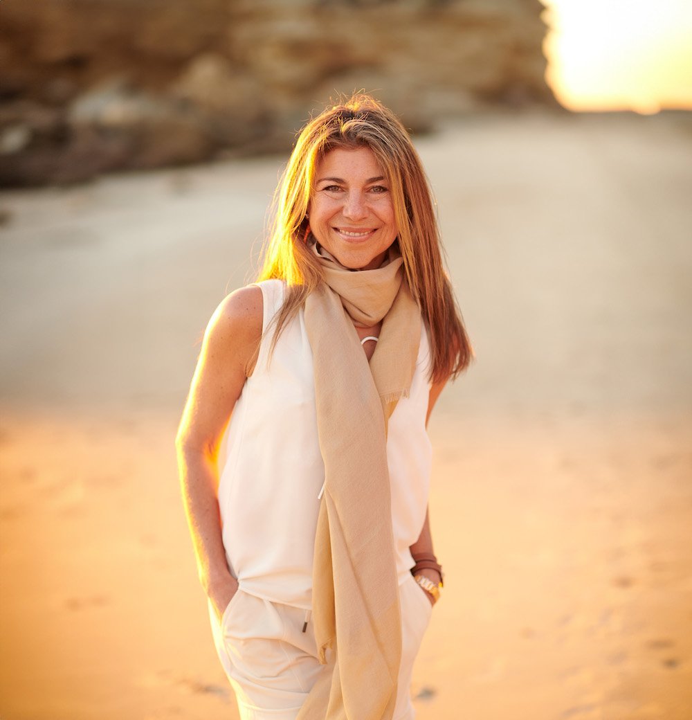 Woman in cream outfit with scarf standing outdoors in natural golden sunlight, smiling warmly
