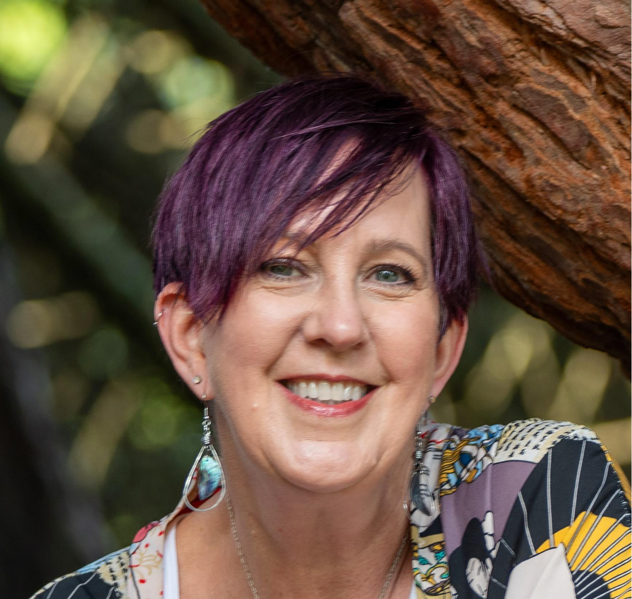 A smiling woman with short purple hair, blue eyes, and earrings, outdoors near a tree with a blurred green background.