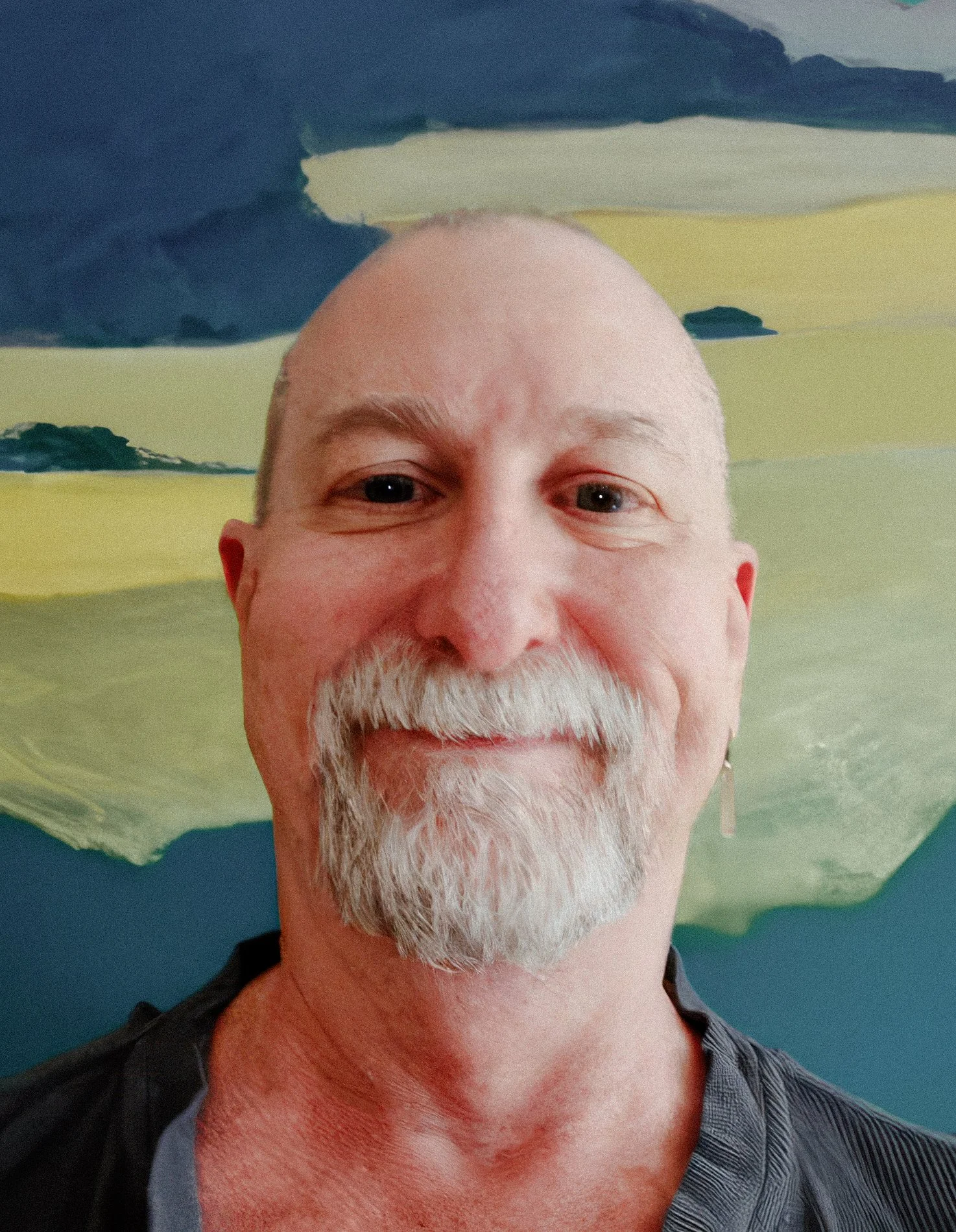 A smiling man with a bald head, gray beard, and mustache, standing in front of an abstract painting with green, yellow, and blue brushstrokes.