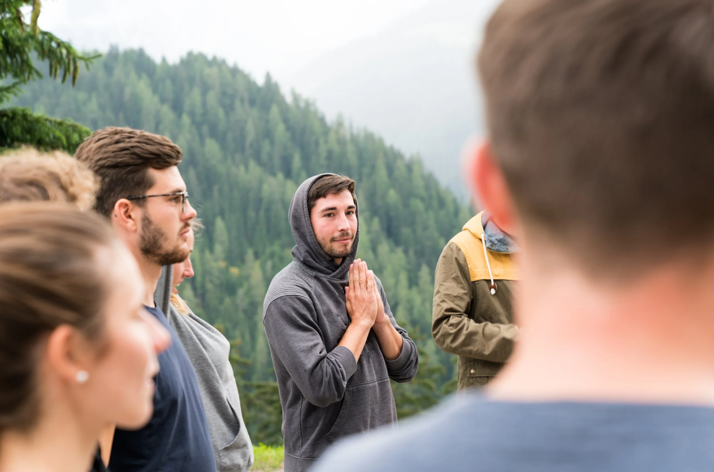 A group of people gathered outdoors in a forested area, with one young man in a gray hoodie and hands together in a prayer or meditation gesture, surrounded by others listening attentively.