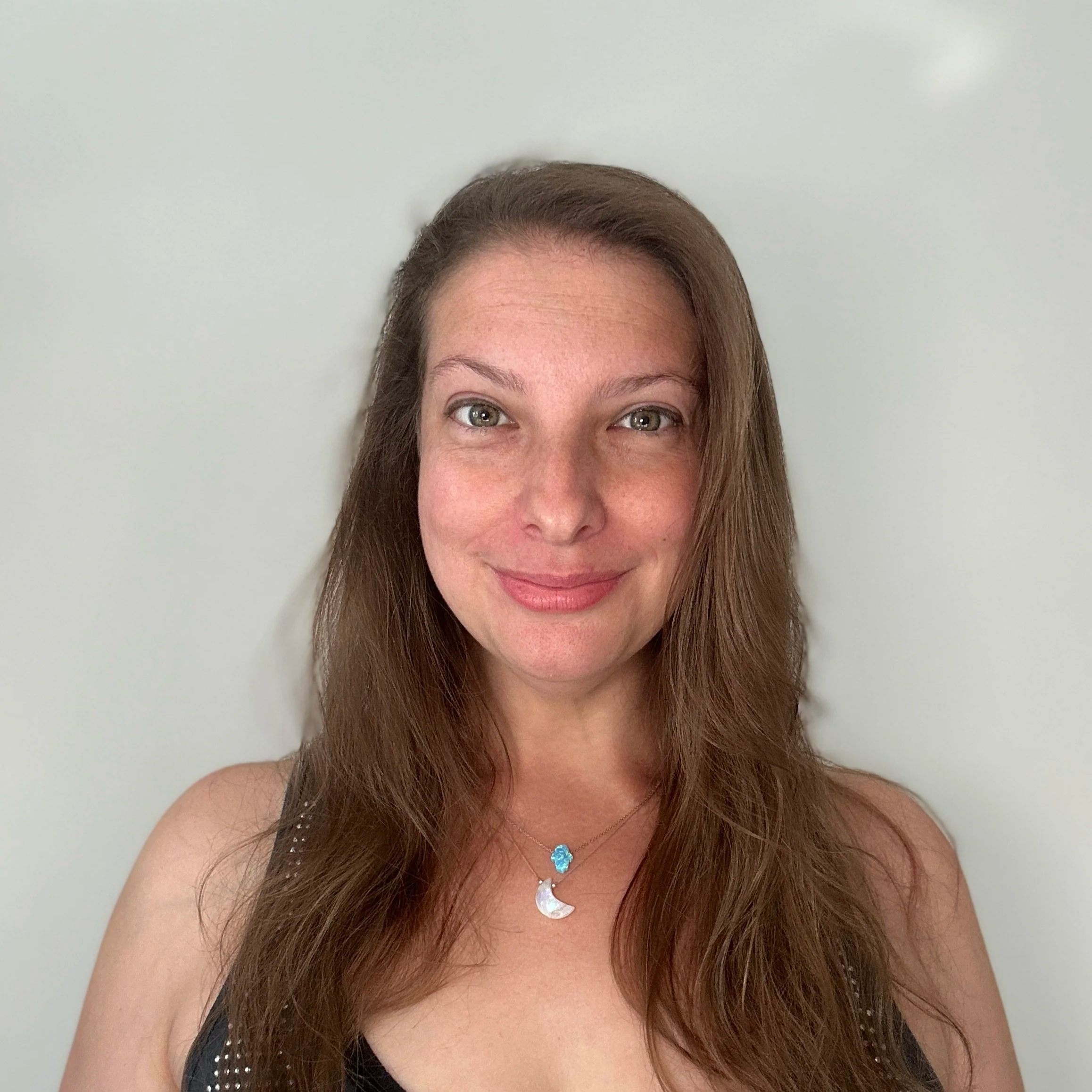 Portrait of a woman with long brown hair, smiling, wearing a black top with metallic details and a moon and star necklace, against a plain light-colored background.