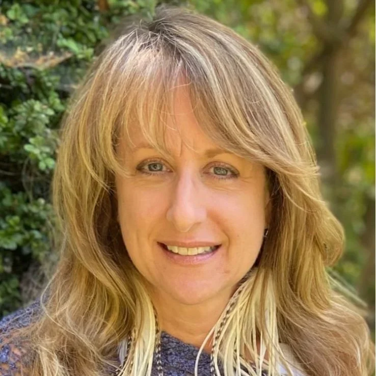 A woman with long, wavy blonde hair smiling outdoors with greenery in the background.