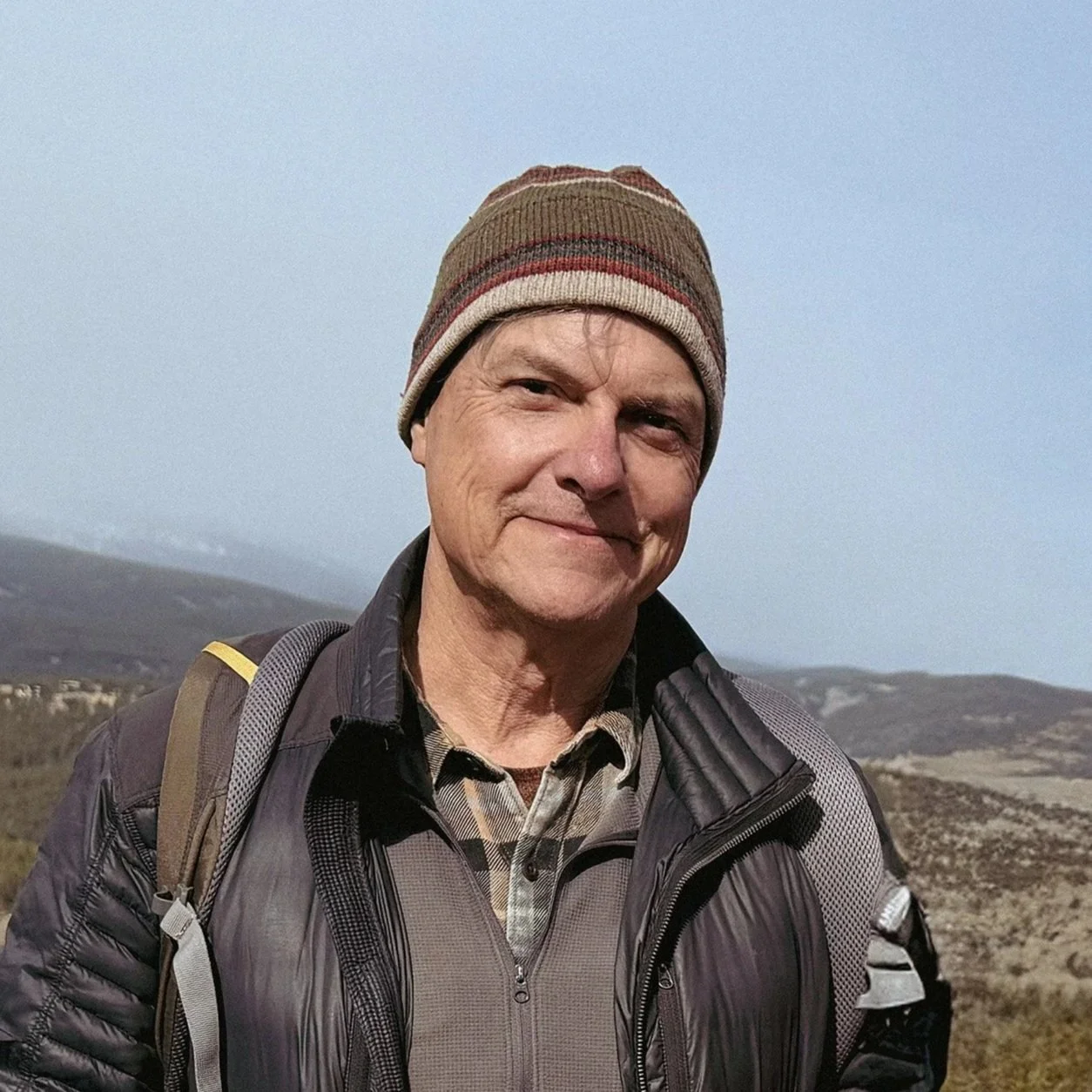 A middle-aged man wearing a striped beanie, a black quilted jacket, and a plaid shirt, standing outdoors in a mountainous landscape with a clear blue sky.