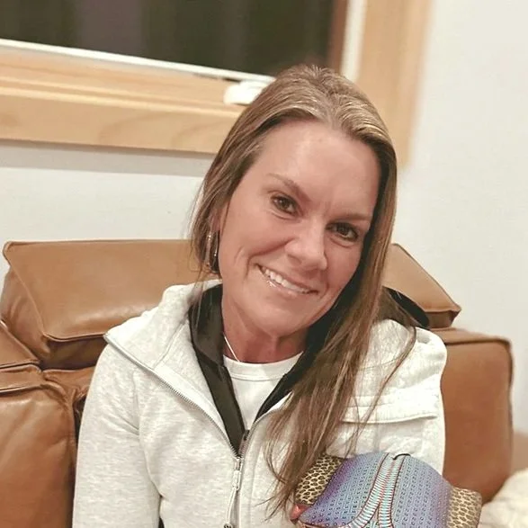 A woman with long brown hair smiling, sitting on a tan leather couch in front of a window.
