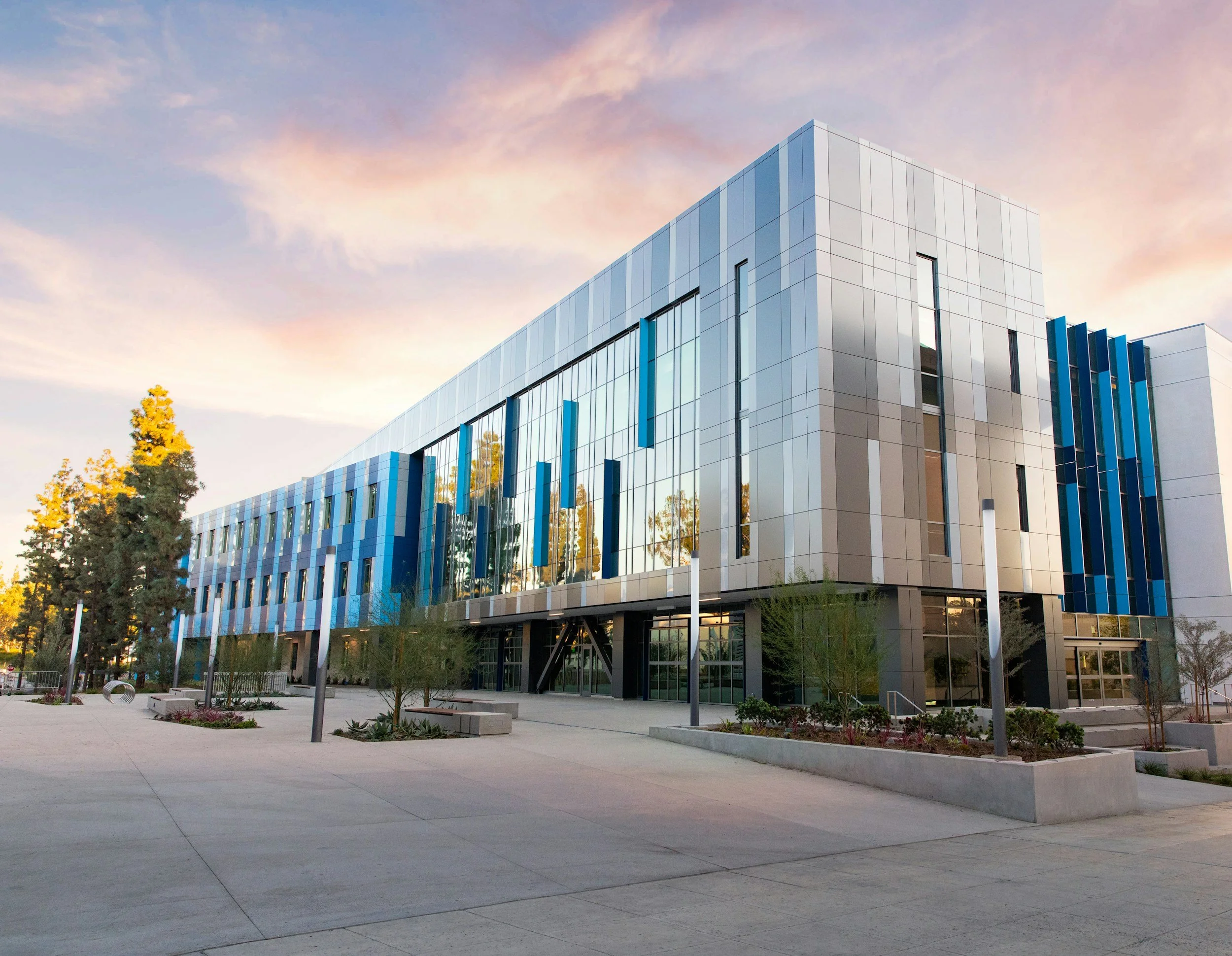 Modern multi-story office building with reflective glass windows and blue vertical accents, surrounded by trees and a paved courtyard during sunset.