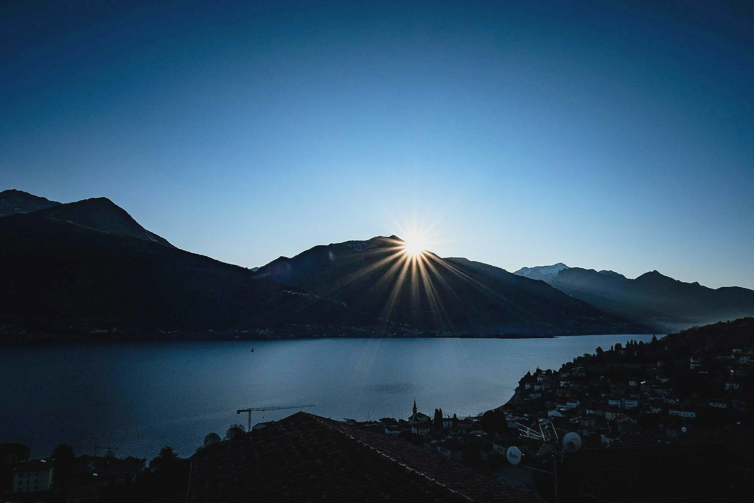 Sun rising behind mountain range over lake, houses and rooftops in foreground.