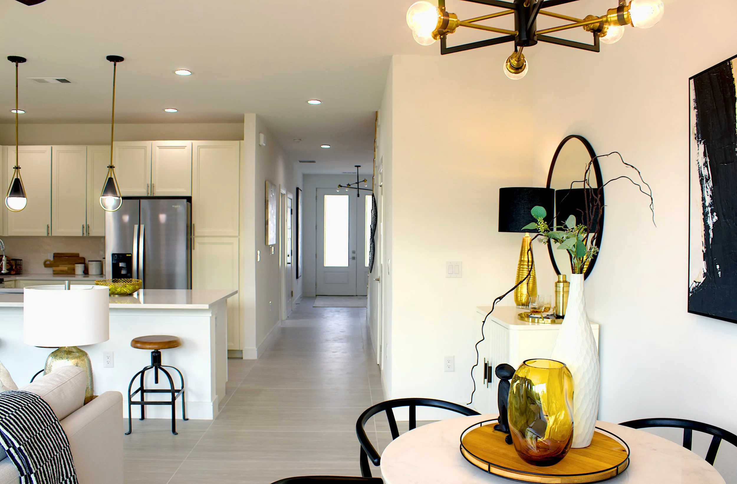 Interior of a modern open-plan home featuring a kitchen with white cabinetry, a dining area with decorative vases and a mirror, and a hallway with artwork and lighting fixtures.