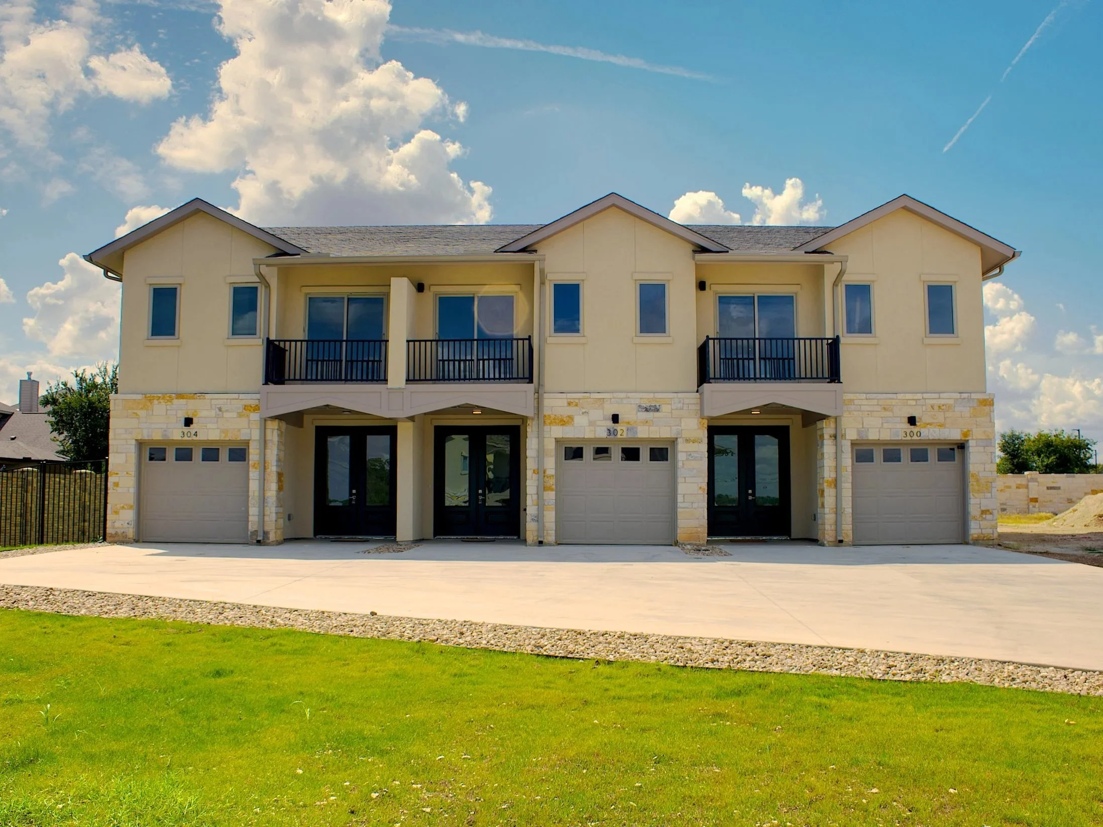 A modern multi-family residential building with three units, each featuring a garage, balcony, and front door, set against a partly cloudy sky.