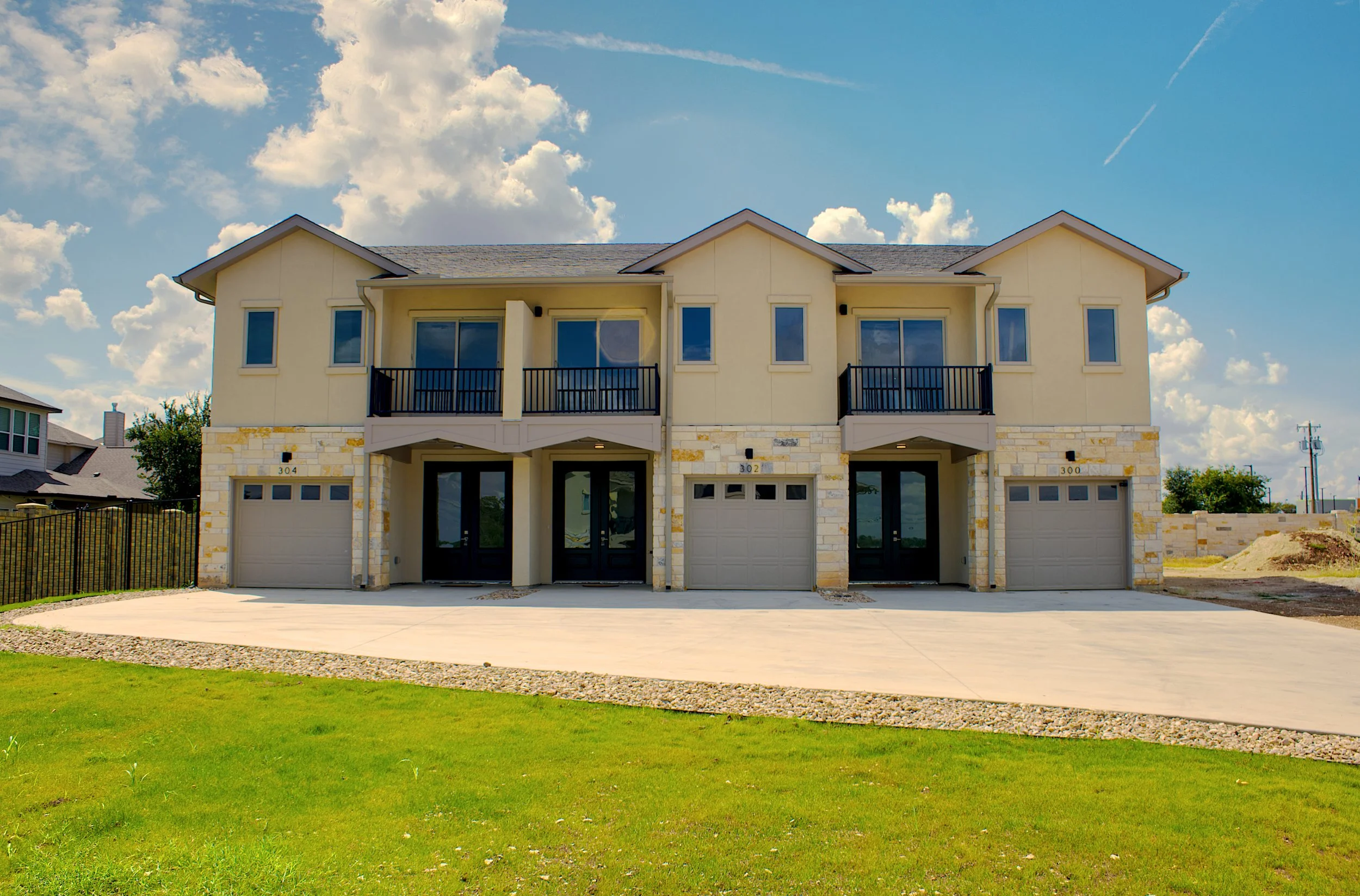 A modern multi-family residential building with three units, each with its own garage and balcony, under a partly cloudy sky.