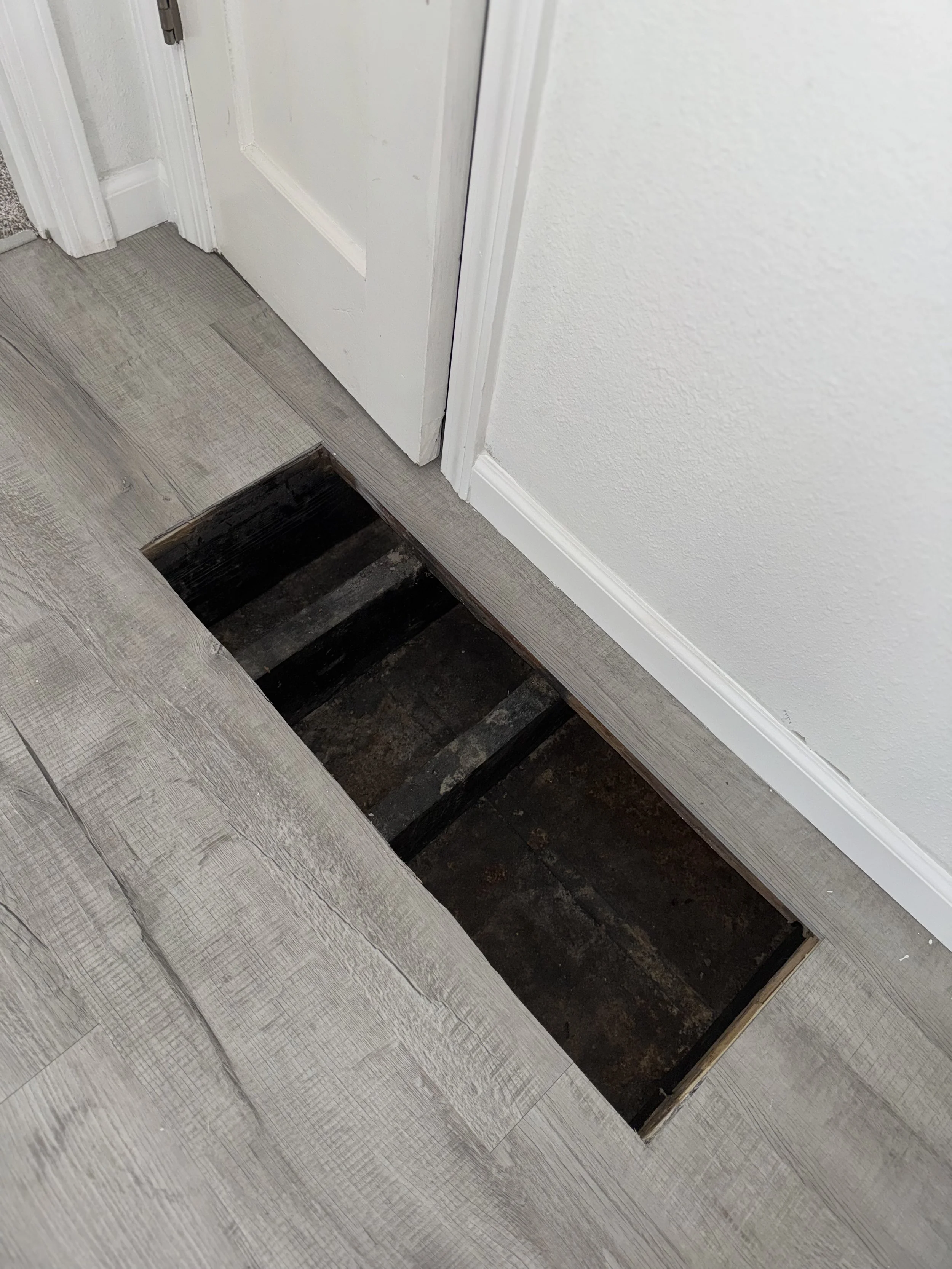 Floor with a large rectangular hole revealing the underlying subfloor and framing, near a white door and baseboard.