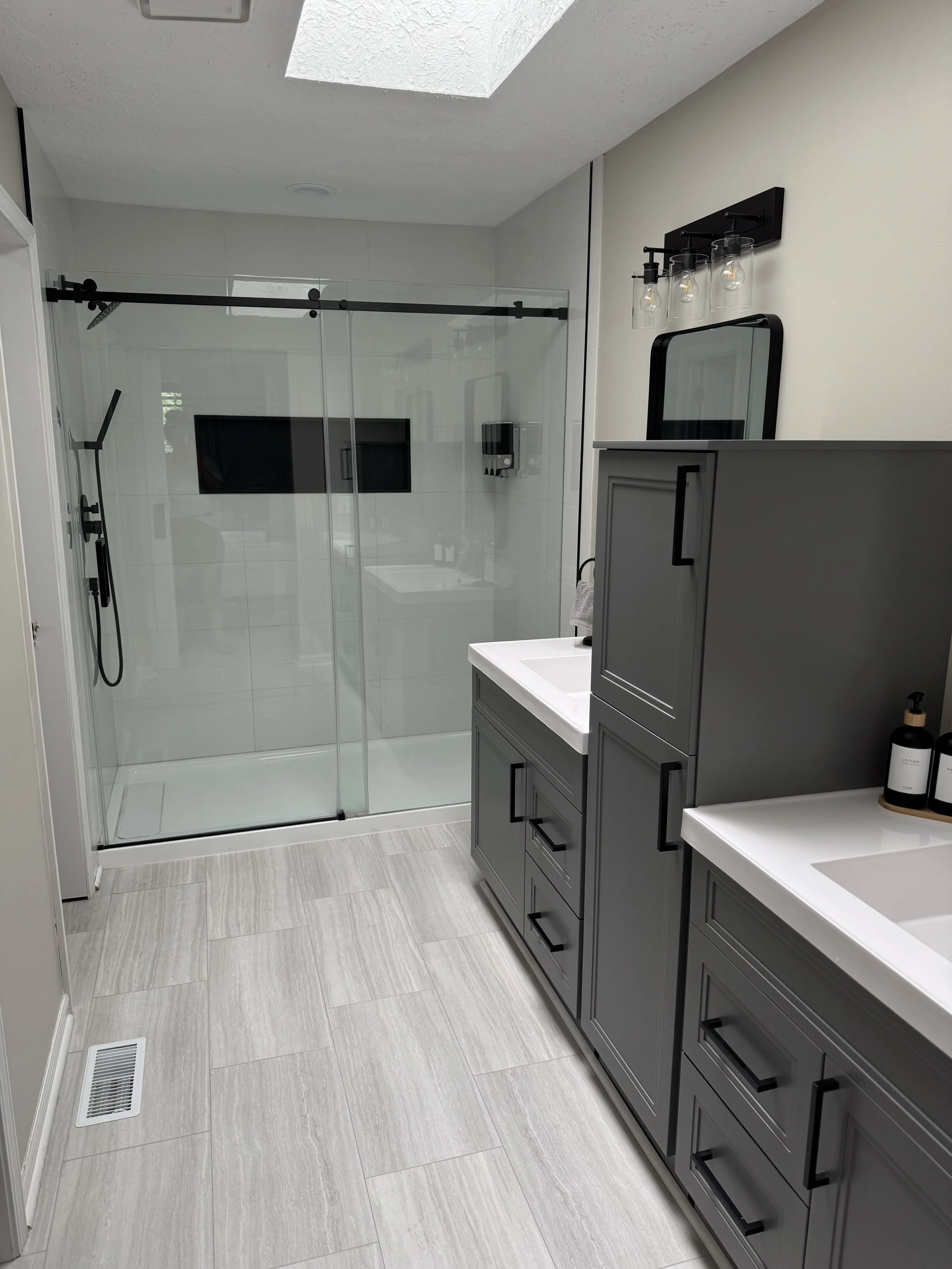 Bathroom with gray double vanity, white sinks, black handles, glass shower enclosure with black hardware, and a wall-mounted mirror and light fixture.