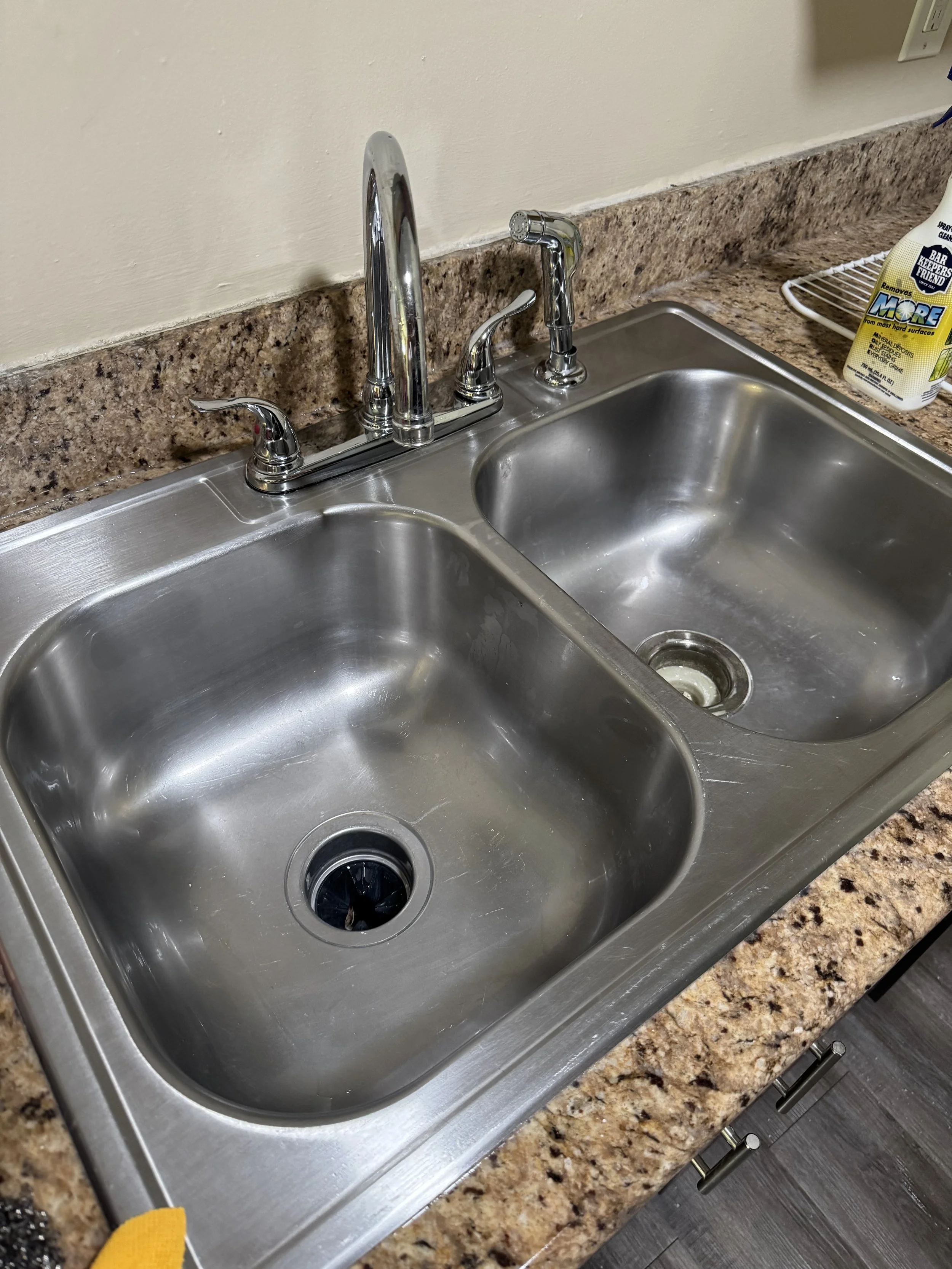 Double stainless steel kitchen sink with chrome faucet and soap dispenser, set into a brown granite countertop, with a bottle of cleaning product on the right and a draining rack in the background.