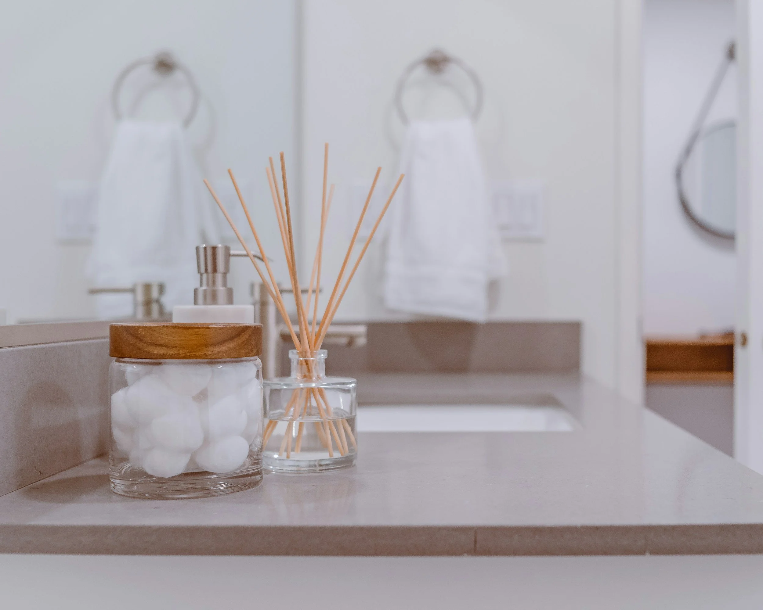 A bathroom countertop with a glass jar of cotton balls, a glass container of reed diffusers, a liquid soap dispenser, and a mirror reflected on a wall.