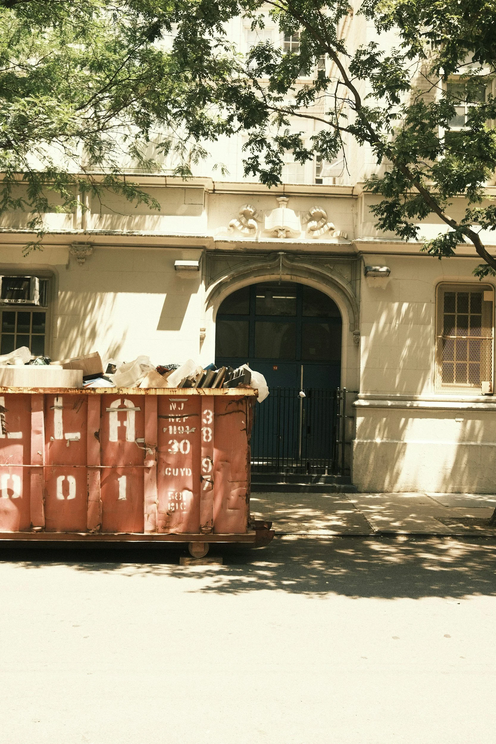 A large dumpster filled with trash is positioned on the sidewalk in front of a beige apartment building. The building has arched windows and decorative architectural details, with trees casting shadows on the facade.