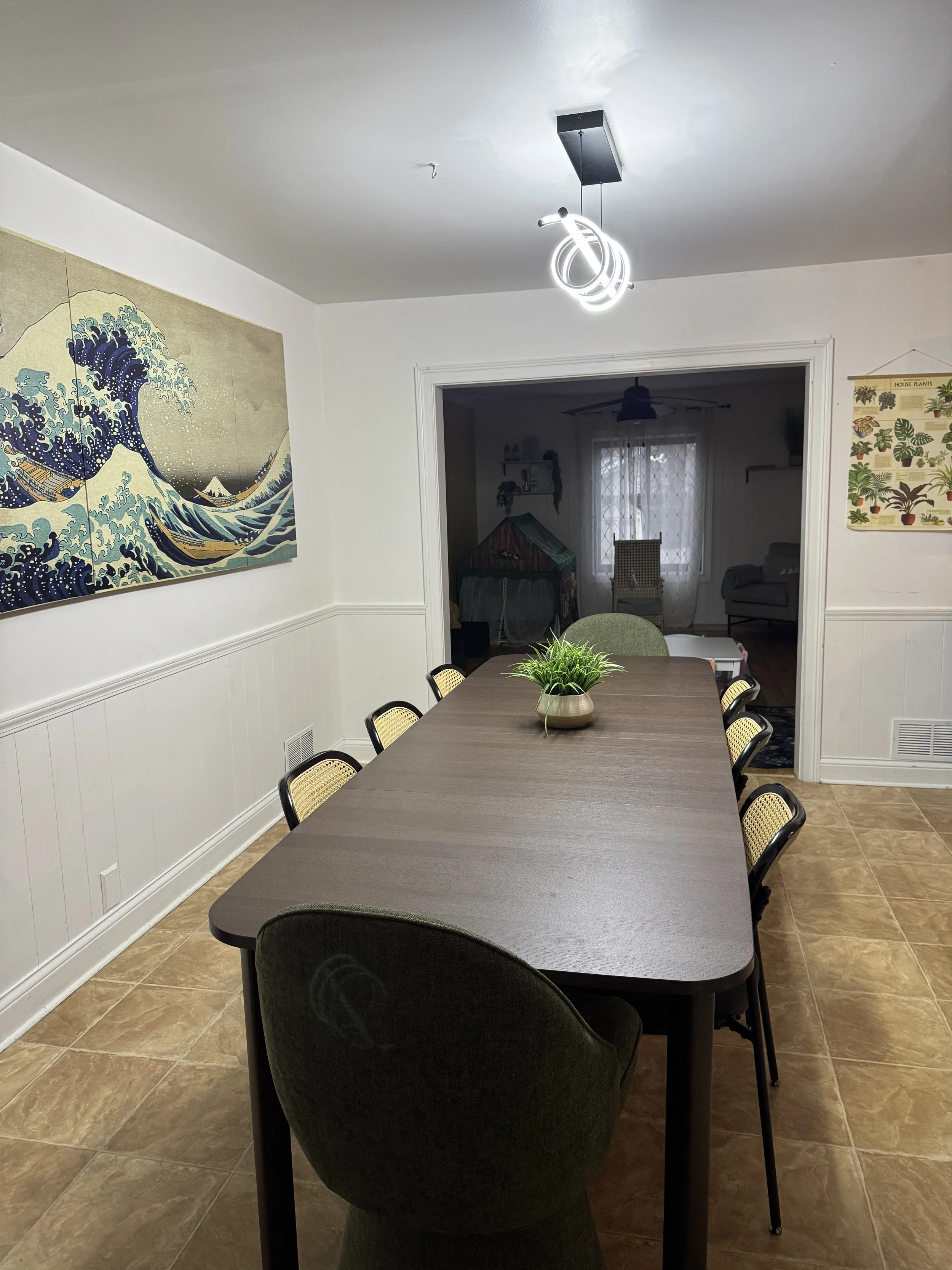 Dining room with a long wooden table, green and black chairs, a plant centerpiece, a modern pendant light, a large wall art piece, and a view into the living room.