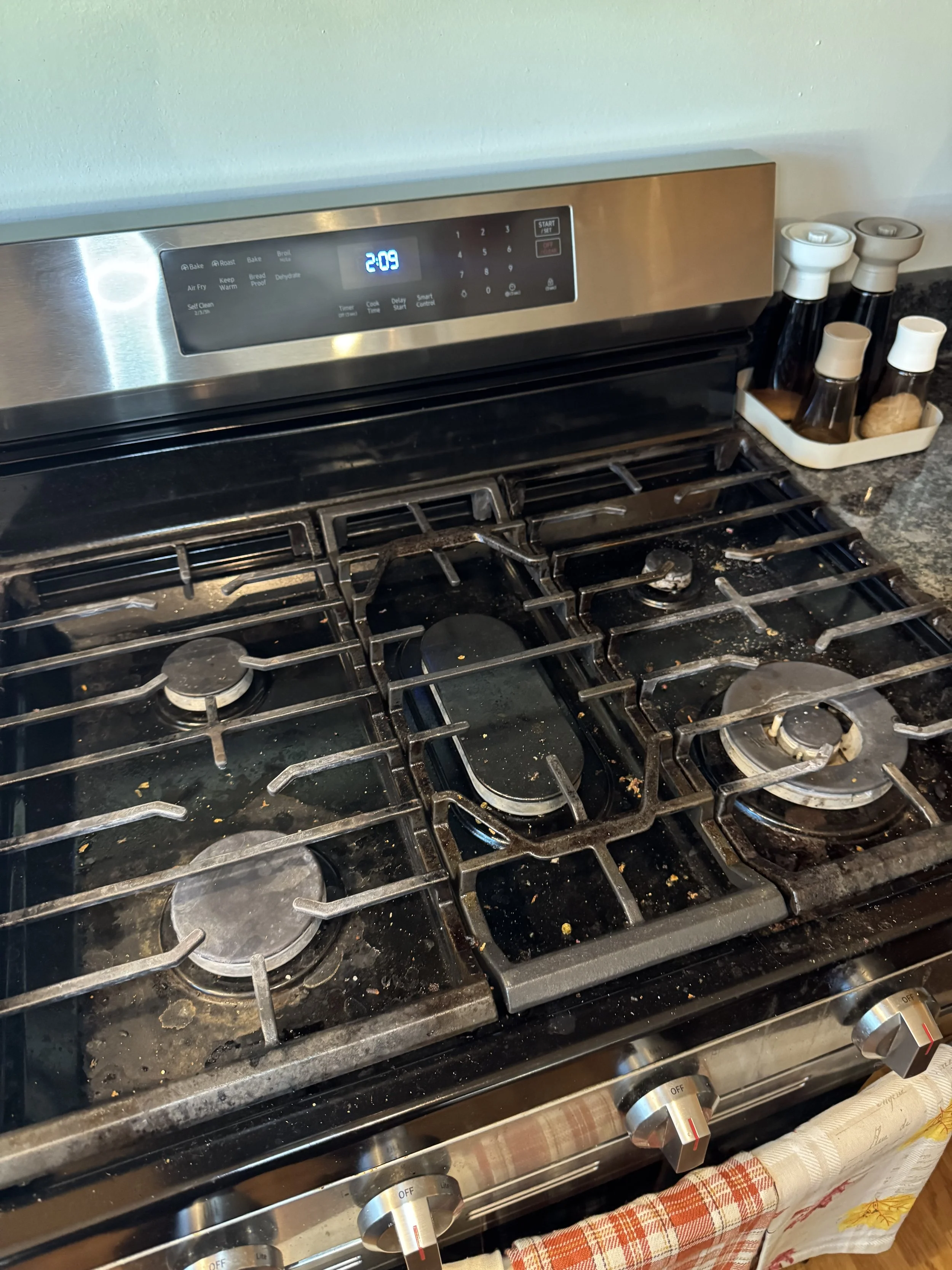 Clean stainless steel gas stove with four burners and a digital clock display reading 2:09. The stovetop has some food crumbs and spills. To the right, there are containers of spices or seasonings in a small tray. The stove has four numbered control knobs.