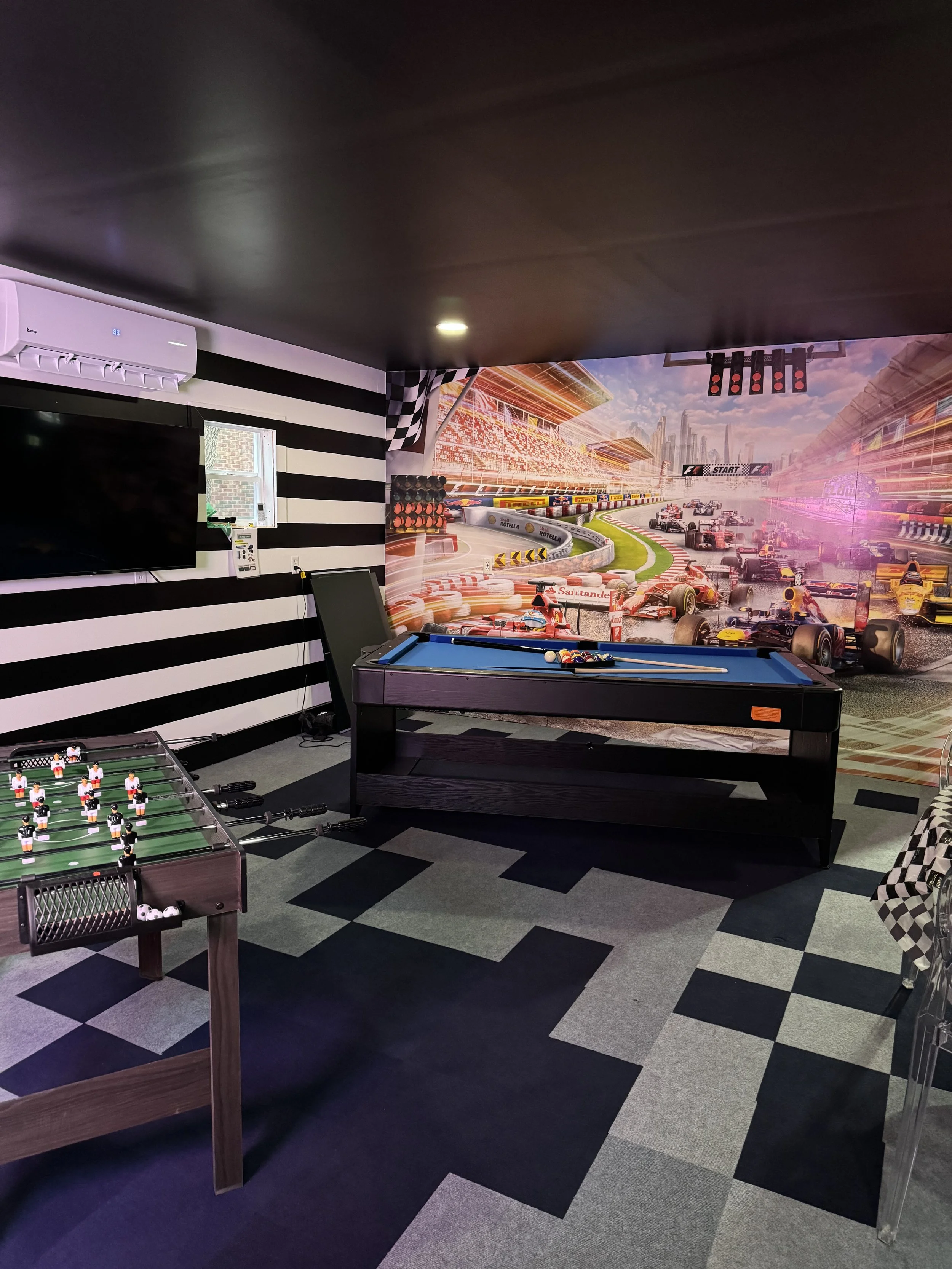 Indoor game room with a mural of a race track featuring race cars, an interactive pool table, Foosball table, and TV mounted on a wall with black and white horizontal stripes.