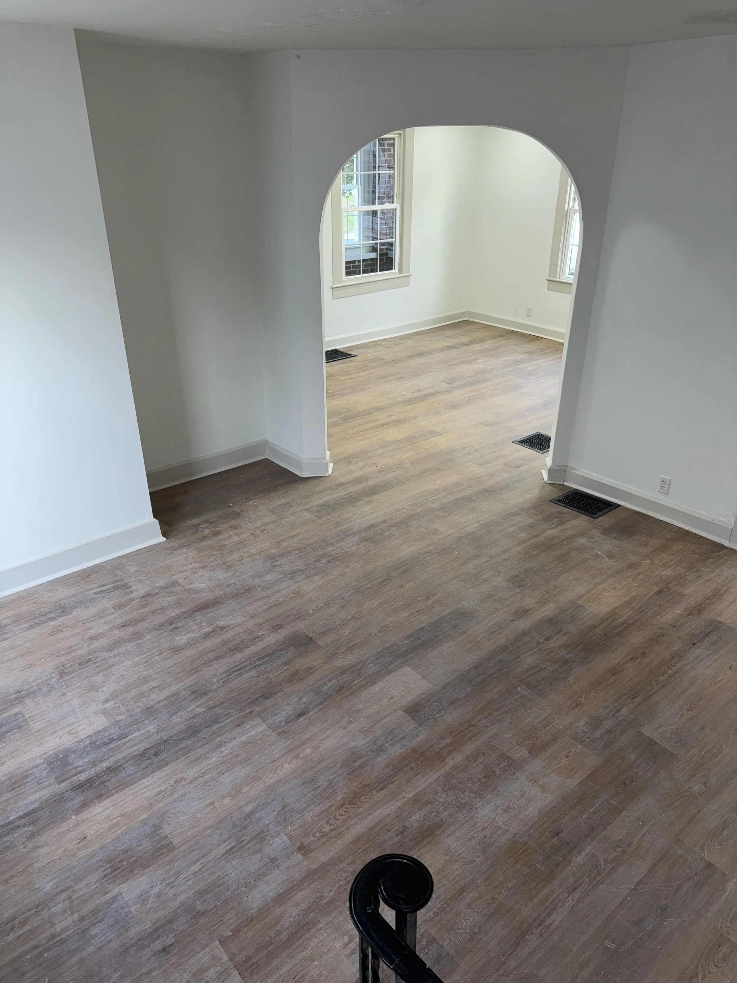 Empty room with hardwood floors, white walls, and multiple windows. An archway leads to an adjacent space, with a black baby gate partially visible at the bottom of the image.