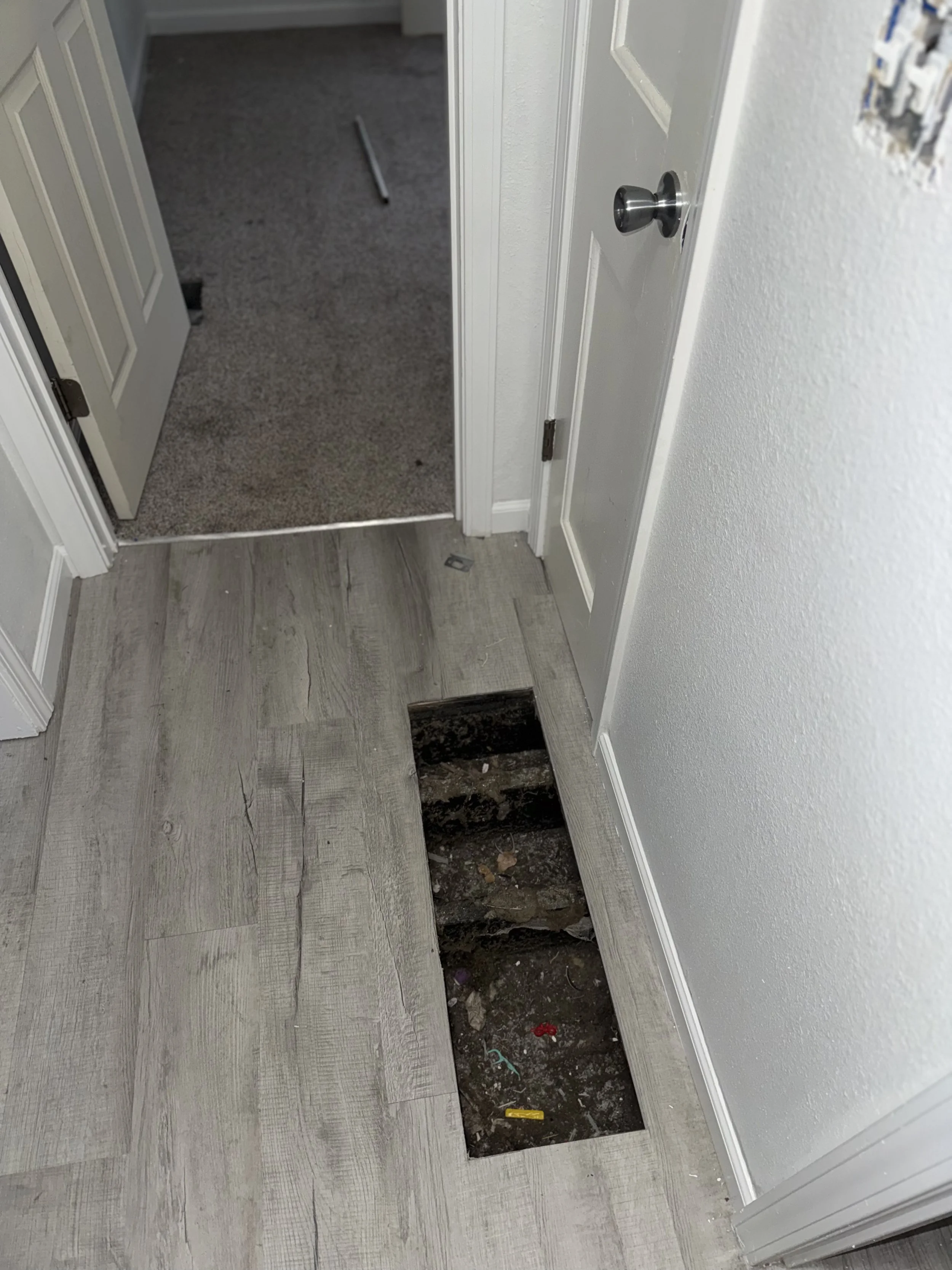 A rectangular hole in the floor reveals plumbing or wiring underneath, next to a closed white door with a silver doorknob in a room with light-colored wood flooring and a wall with a damaged electrical outlet cover.